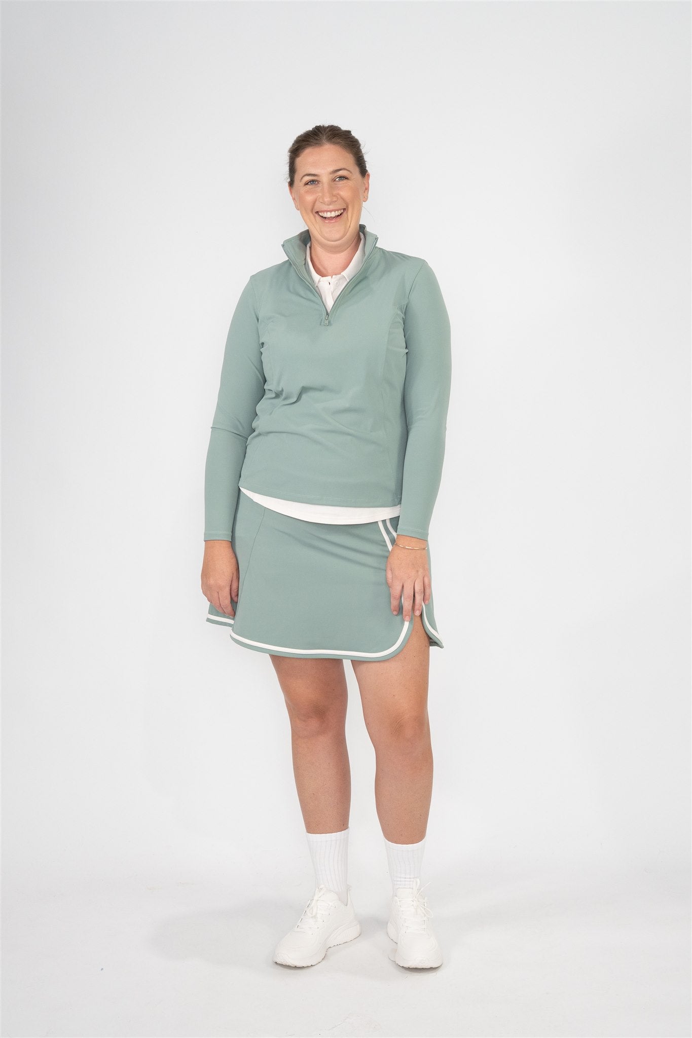Mid-size woman wearing Fairway Green quarter-zip pullover layered over a Sand polo and matching Fairway Green wrap skort with white trim, smiling in a relaxed front-facing pose. Inclusive women’s golf apparel crafted for comfort, coverage, and on-course style with effortless layering.