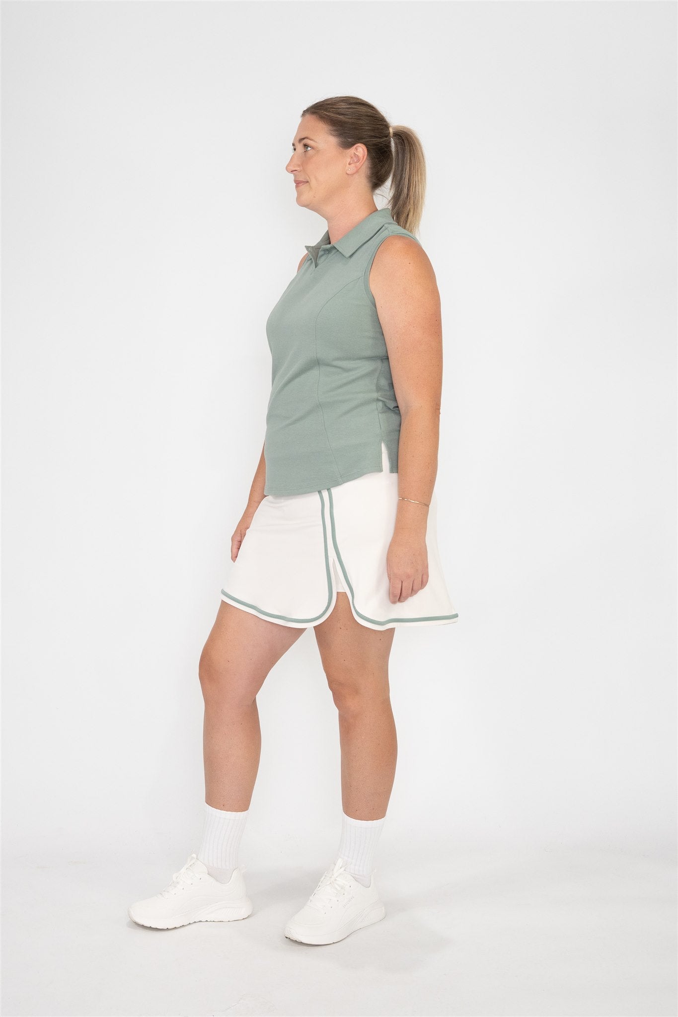 Woman with plus or mid size body wearing fairway green sleeveless golf polo and sand golf skort with fairway green trim, standing in side profile against white background. Stylish and functional women’s golf apparel designed for fashionable and active players of all sizes.