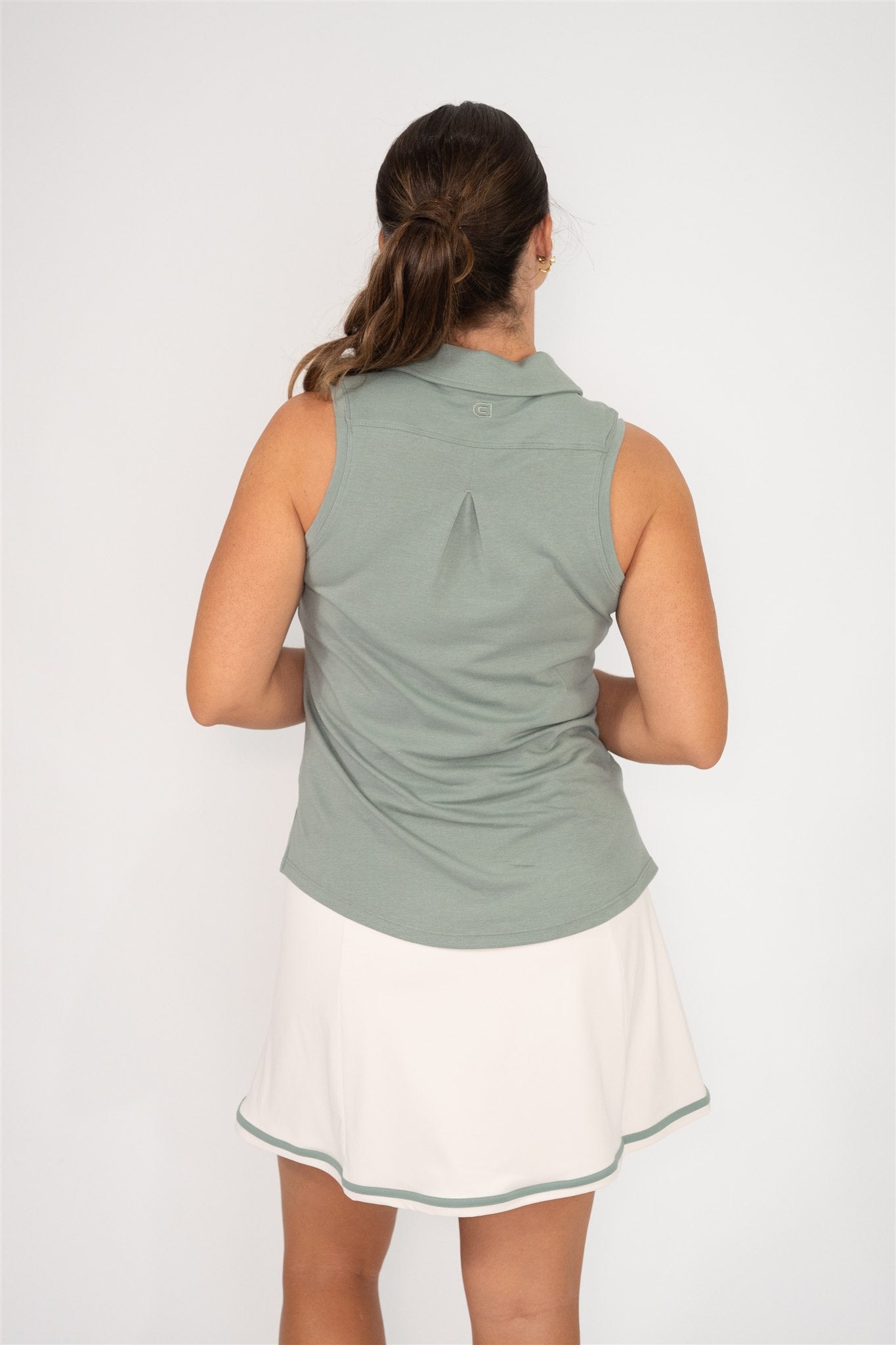 Woman wearing fairway green sleeveless golf polo and sand golf skort with fairway green trim, shown from back with hair in ponytail, highlighting back pleat detail and relaxed fit against white background. Elegant and functional women’s golf apparel for modern players.