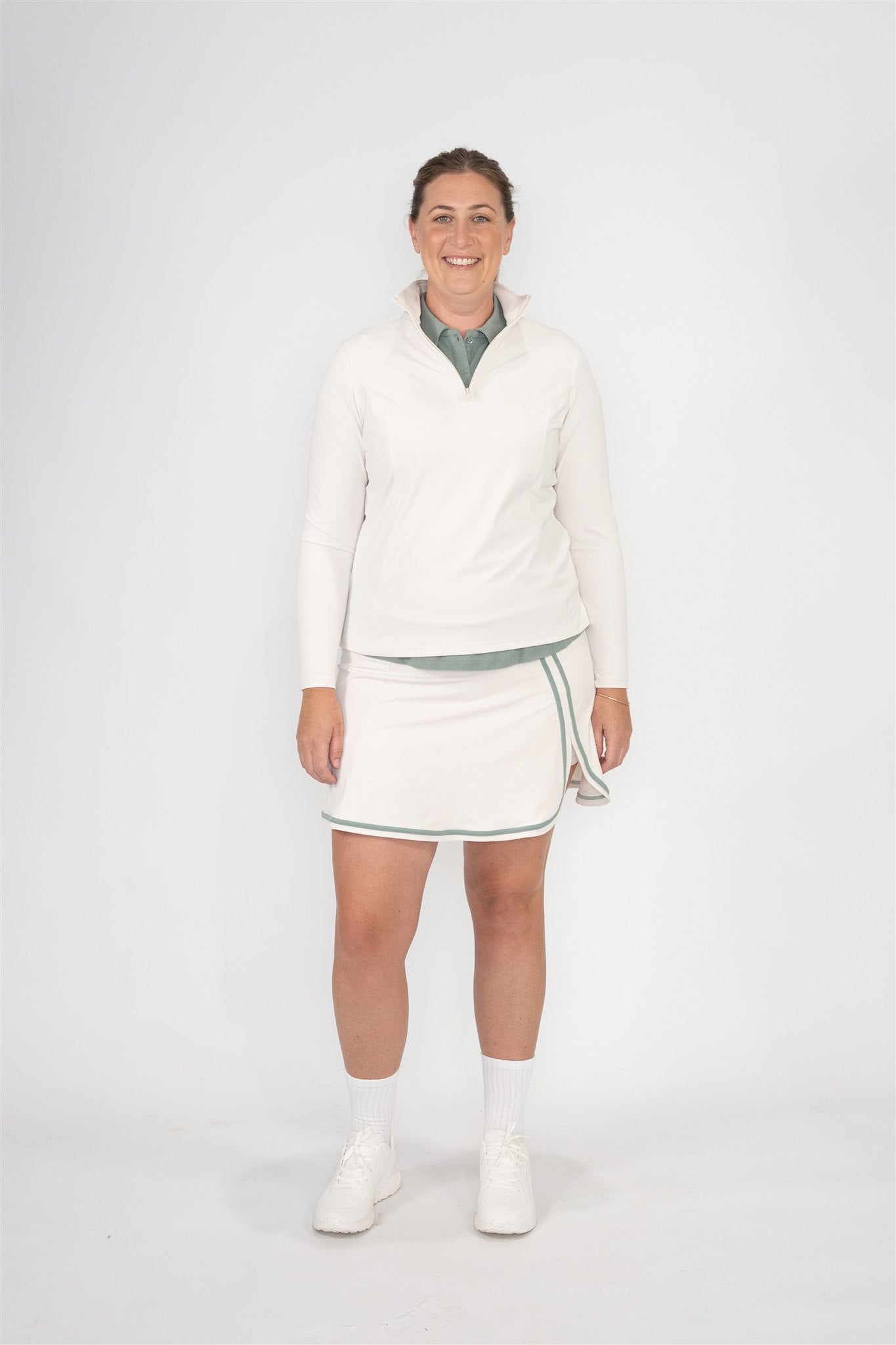 Mid-size woman wearing Sand quarter-zip pullover layered over a Fairway Green polo and Sand wrap skort with Fairway Green trim, standing front-facing and smiling. Comfortable and inclusive women’s golf apparel styled for adaptable layering and confident course performance.