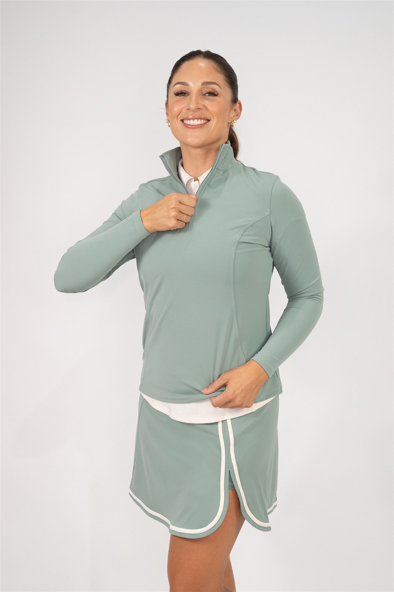 Woman wearing Fairway Green quarter-zip pullover layered over a Sand polo and matching Fairway Green wrap skort with white trim, smiling while adjusting the zipper. Comfortable and stylish women’s golf apparel crafted for performance, movement, and layering versatility.