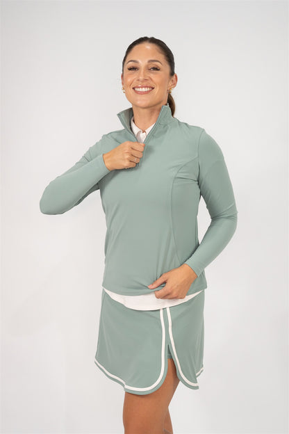 Woman wearing Fairway Green quarter-zip pullover layered over a Sand polo and matching Fairway Green wrap skort with white trim, smiling while adjusting the zipper. Comfortable and stylish women’s golf apparel crafted for performance, movement, and layering versatility.