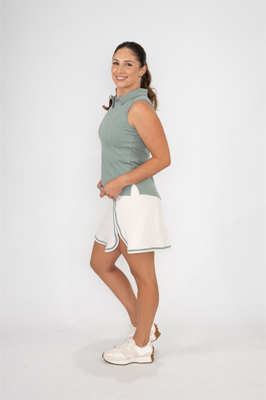 Woman wearing fairway green sleeveless golf polo and sand golf skort with fairway green trim, smiling and posing sideways against white background. Elegant and functional women’s golf apparel for stylish and active players.