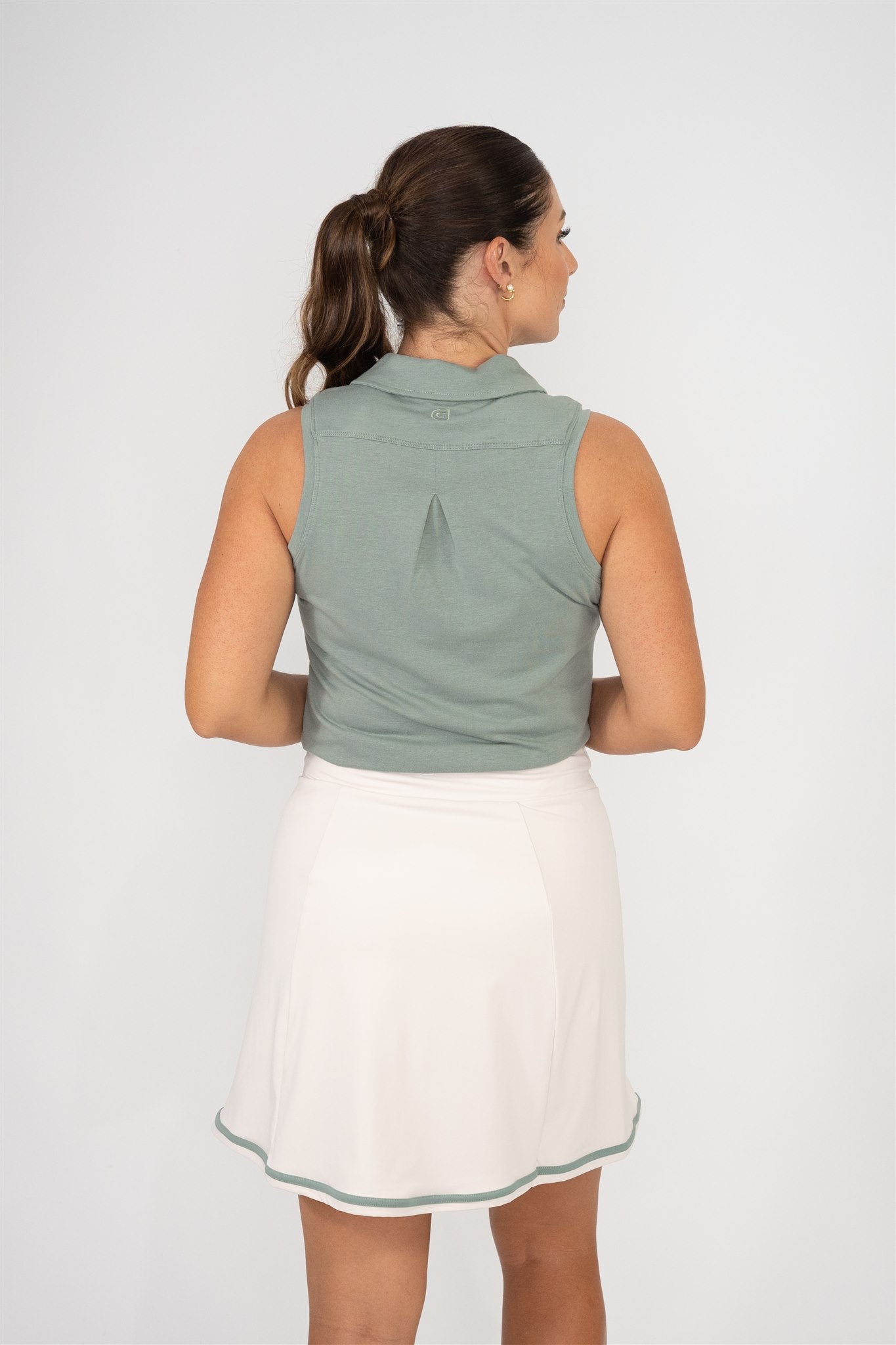 Woman wearing fairway green sleeveless golf polo and sand golf skort with fairway green trim, shown from back with hair in ponytail, highlighting back pleat and skort design against white background. Elegant and functional women’s golf apparel for modern players.