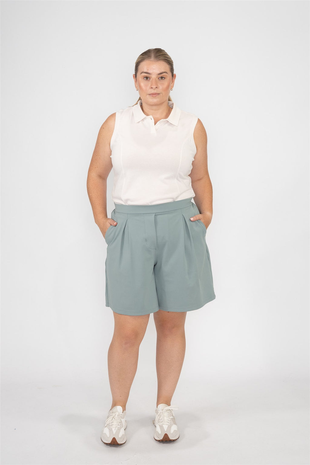 Mid-sized woman wearing Sand sleeveless polo and Fairway Green pleated shorts, standing front-facing against a studio background. Sophisticated and easygoing women’s golf apparel designed for all-day comfort and movement.