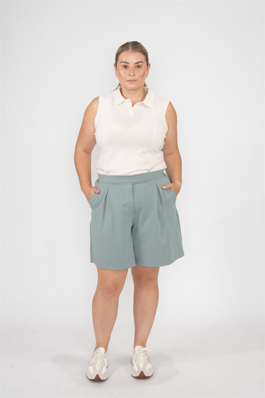 Mid-sized woman wearing Sand sleeveless polo and Fairway Green pleated shorts, standing front-facing against a studio background. Sophisticated and easygoing women’s golf apparel designed for all-day comfort and movement.
