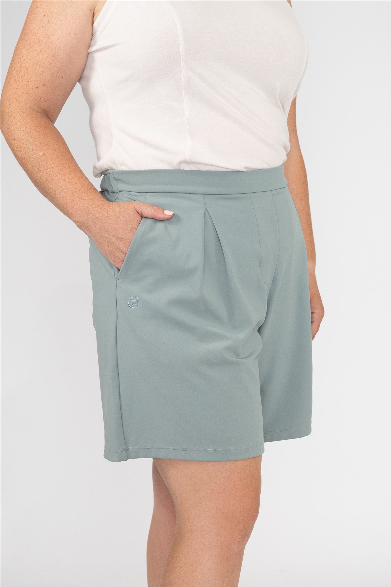 Close-up of mid-sized woman wearing Sand sleeveless polo and Fairway Green pleated golf shorts, with hand in pocket showcasing side pleats and tonal embroidered logo. Elegant and functional detail in women’s golf wear for movement and style.