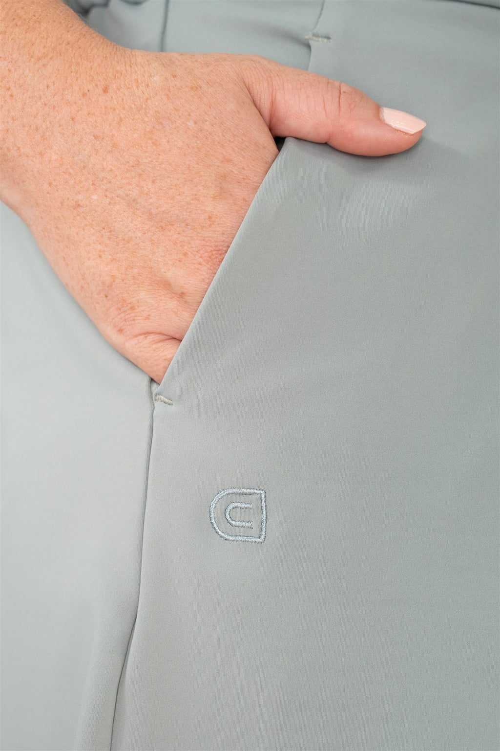 Close-up of mid-sized woman’s hand in pocket of Fairway Green pleated golf shorts, featuring tonal Clubdayze “C” logo embroidery. Premium women’s golf wear designed for function, detail, and elegant movement.