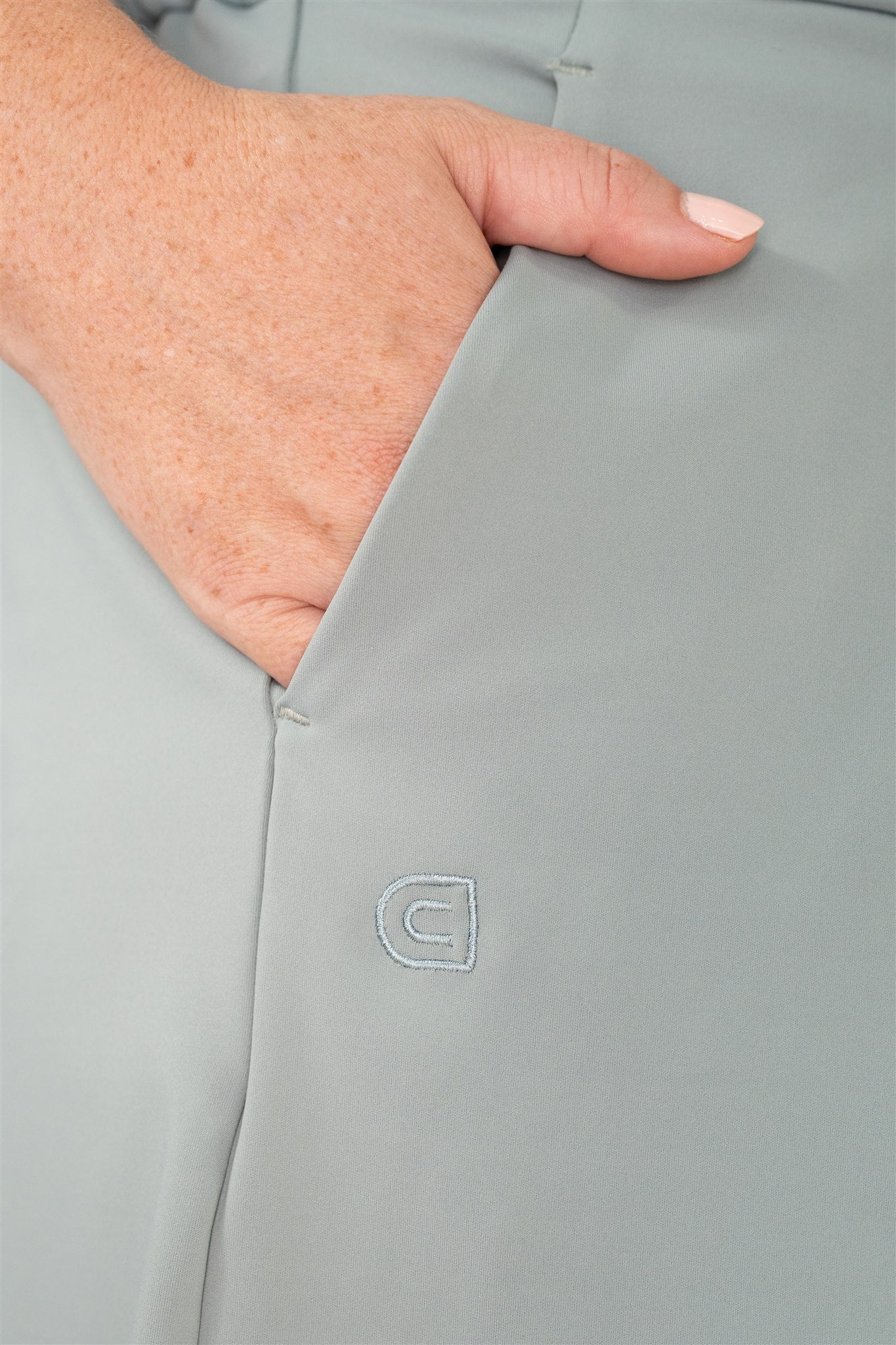 Close-up of mid-sized woman’s hand in pocket of Fairway Green pleated golf shorts, featuring tonal Clubdayze “C” logo embroidery. Premium women’s golf wear designed for function, detail, and elegant movement.