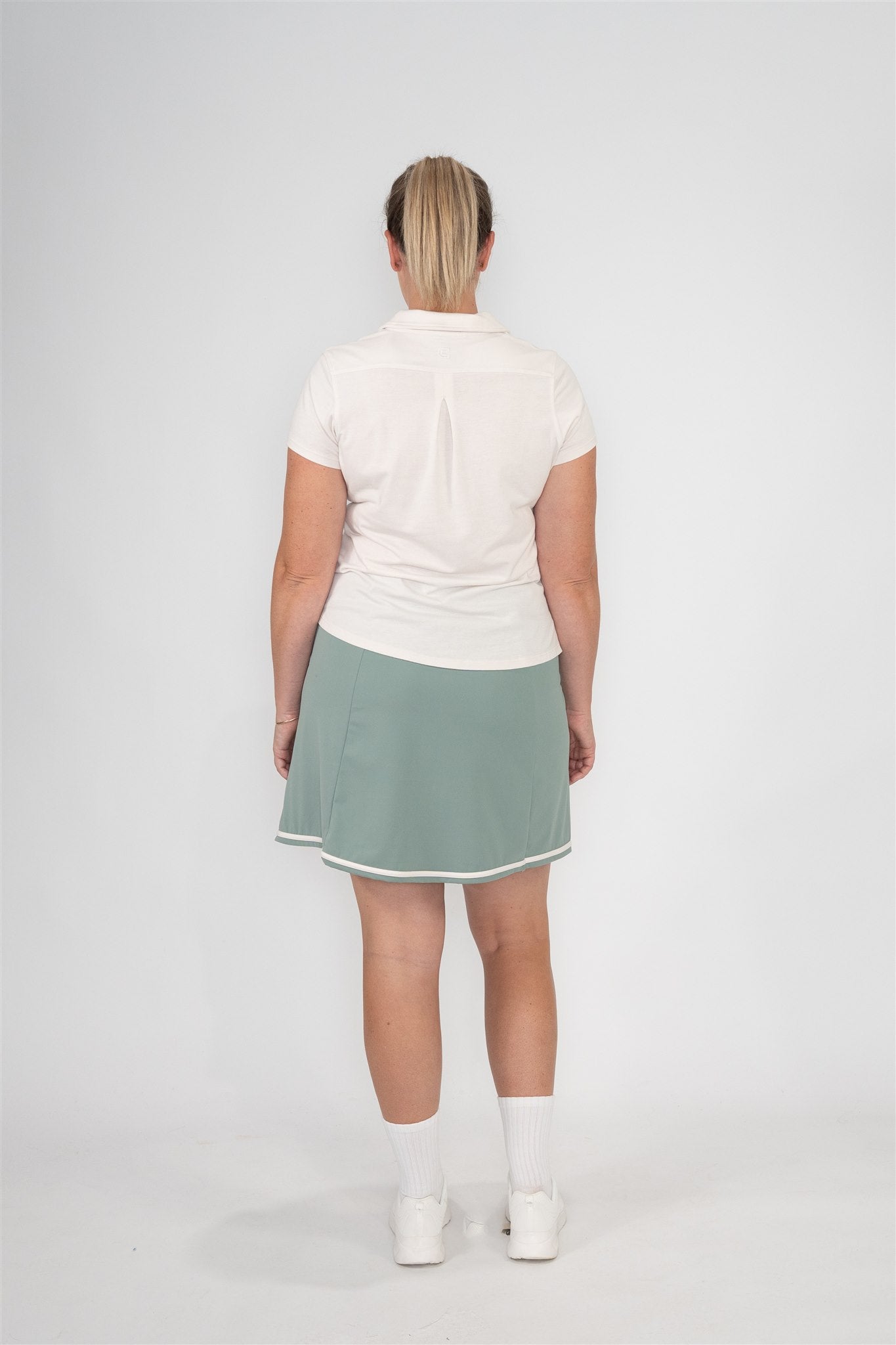 Mid-size woman wearing Sand short sleeve polo and Fairway Green wrap skort with white trim, shown from the back against a clean studio background. Flattering and functional women’s golf apparel offering breathable comfort and inclusive fit for active lifestyles.
