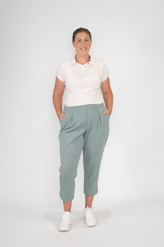 Mid-size woman wearing Sand short sleeve polo and Fairway Green cropped golf trousers with front pleats, standing front-facing with hands in pockets against a studio backdrop. Refined and inclusive women’s golf apparel designed for relaxed elegance and all-day performance.