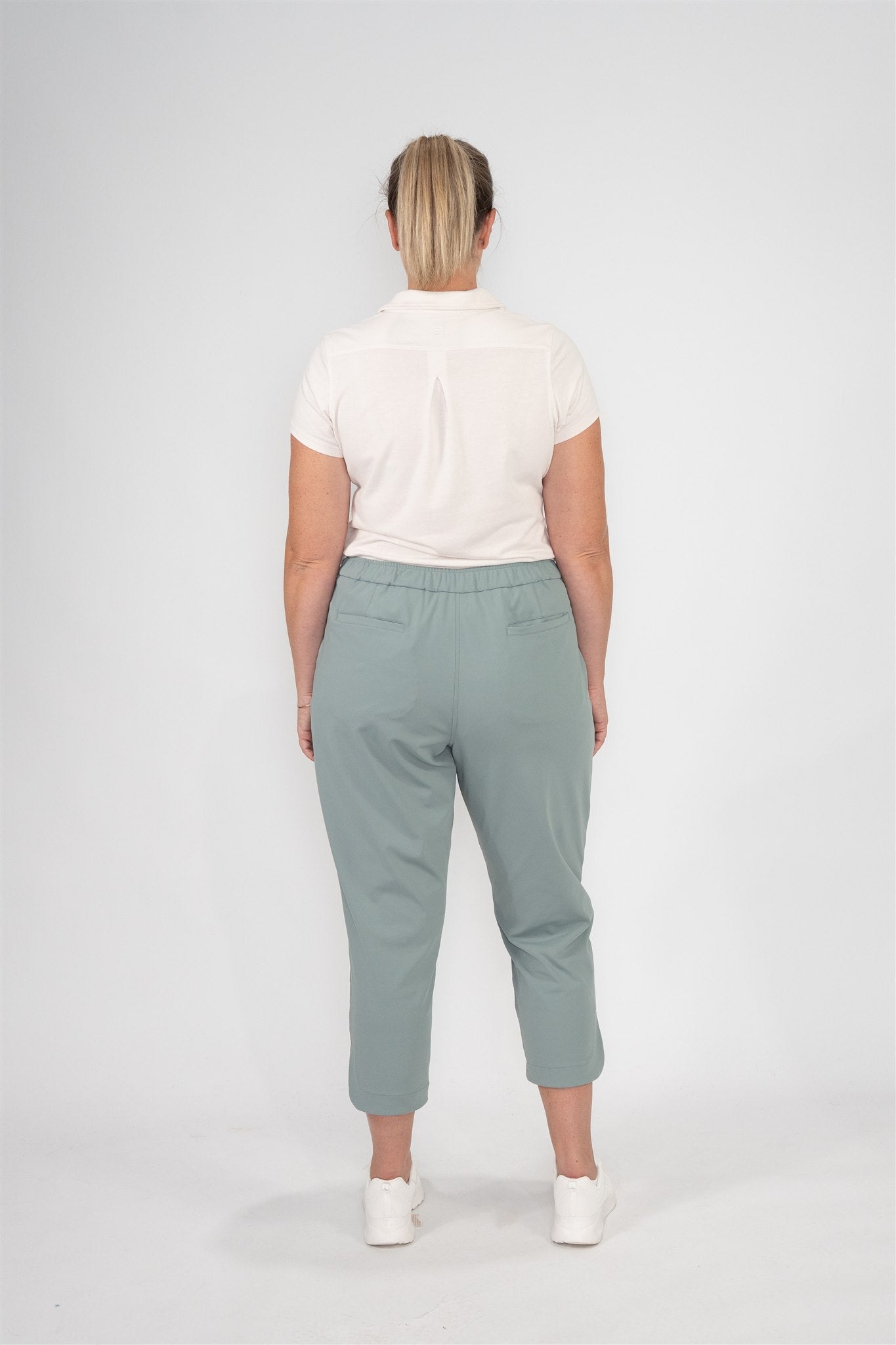 Mid-sized woman wearing Sand short-sleeve polo and Fairway Green tapered ankle golf pants, back view showing elastic waistband and welt pockets. Clean white studio setting. Elegant and functional women’s golf apparel for stylish movement and all-day comfort.
