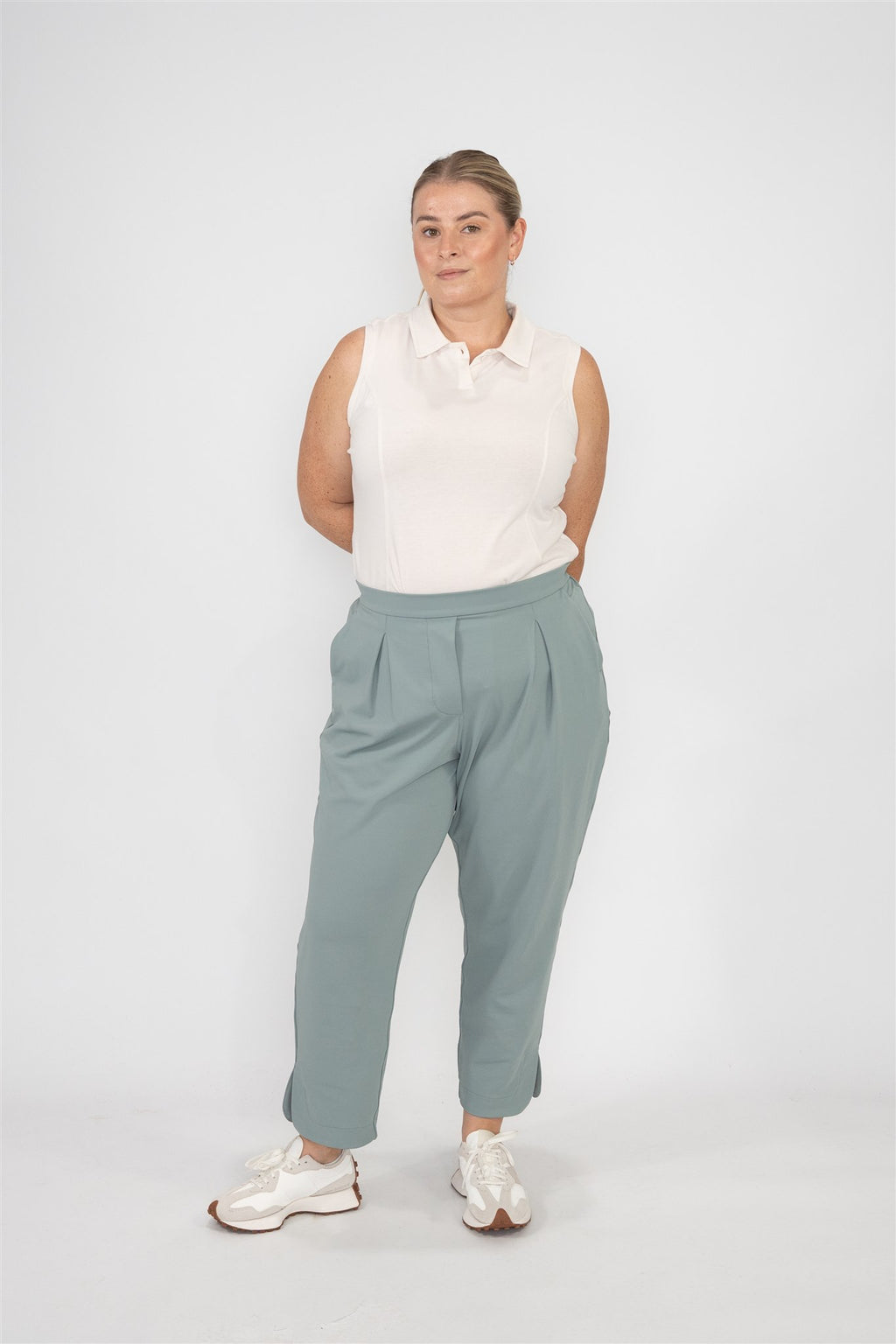 Mid-sized woman wearing Sand sleeveless polo and Fairway Green tapered ankle pants, standing with hands behind back in a relaxed front-facing pose against a white studio backdrop. Elegant and versatile women’s golf apparel for confident, all-day wear on and off the course.
