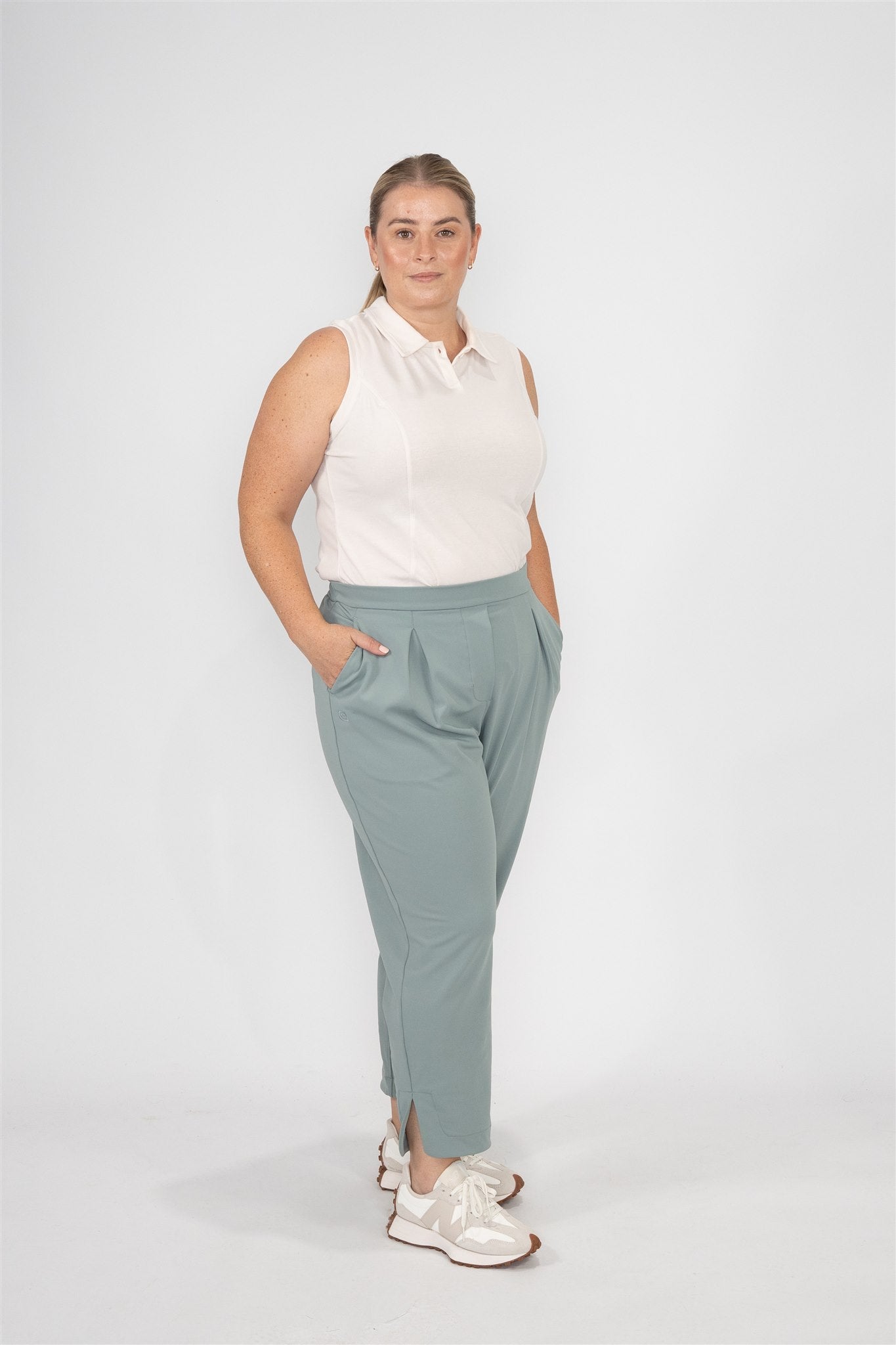 Mid-sized woman wearing Sand sleeveless polo and Fairway Green tapered ankle pants with pleats, hands in pockets, standing in a soft front-facing pose against a white studio background. Effortless and flattering women’s golf apparel crafted for movement and modern elegance.