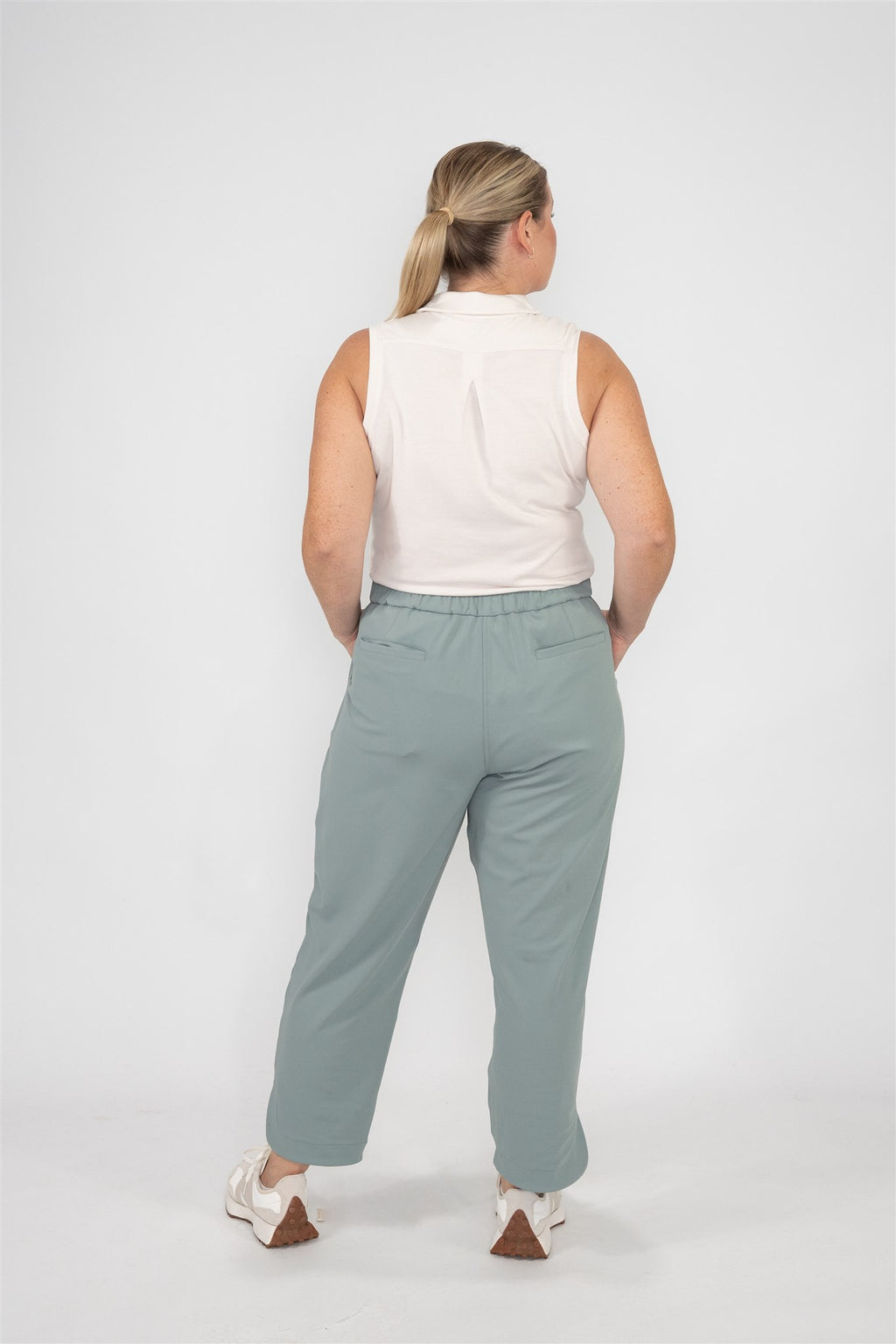 Mid-sized woman wearing Sand sleeveless polo and Fairway Green tapered ankle pants, standing in a relaxed back-facing pose with hands resting on hips against a white studio backdrop. Clean-lined, functional women’s golf wear designed for confidence and comfort on the course.