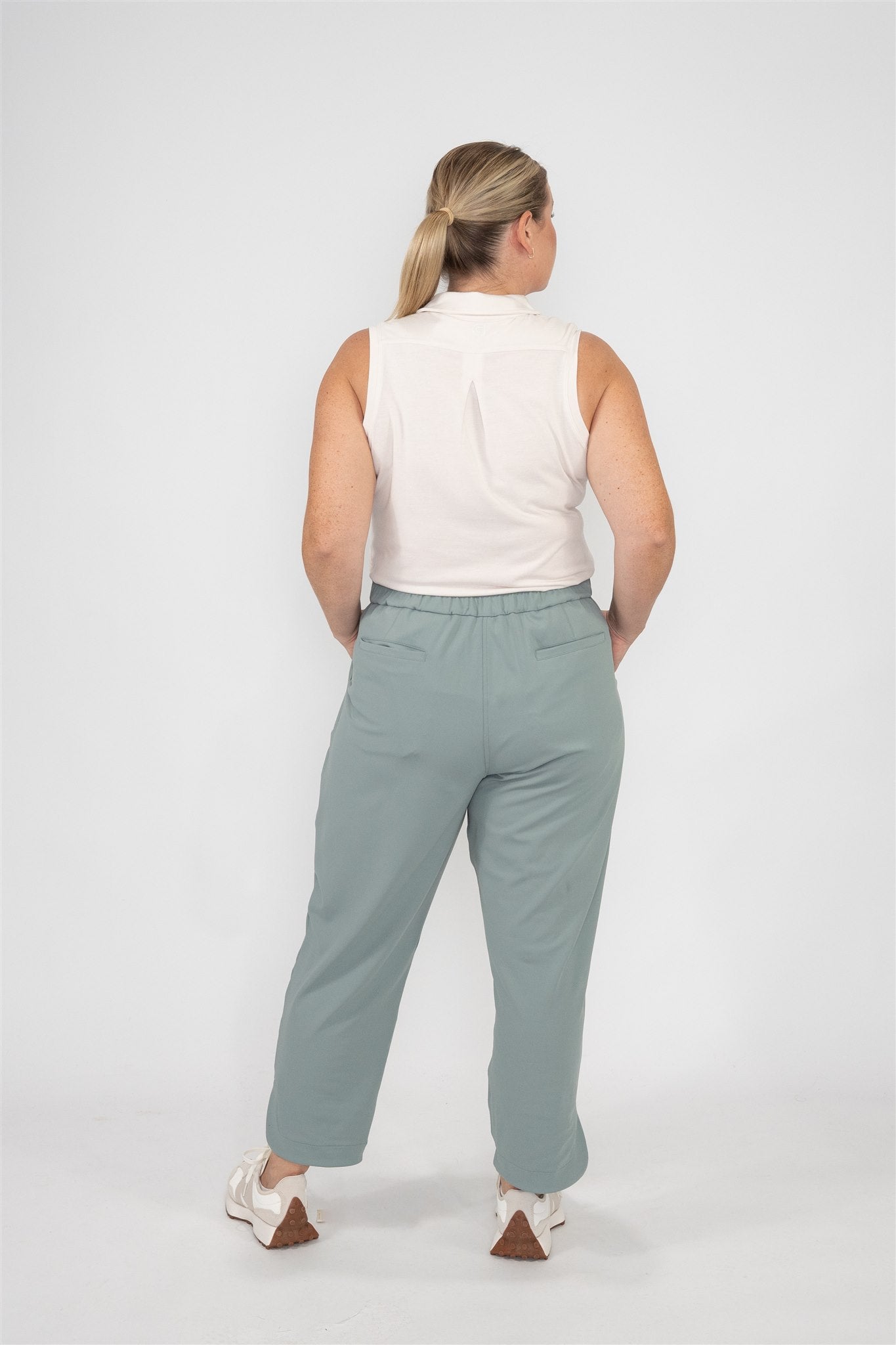 Mid-sized woman wearing Sand sleeveless polo and Fairway Green tapered ankle pants, standing in a relaxed back-facing pose with hands resting on hips against a white studio backdrop. Clean-lined, functional women’s golf wear designed for confidence and comfort on the course.