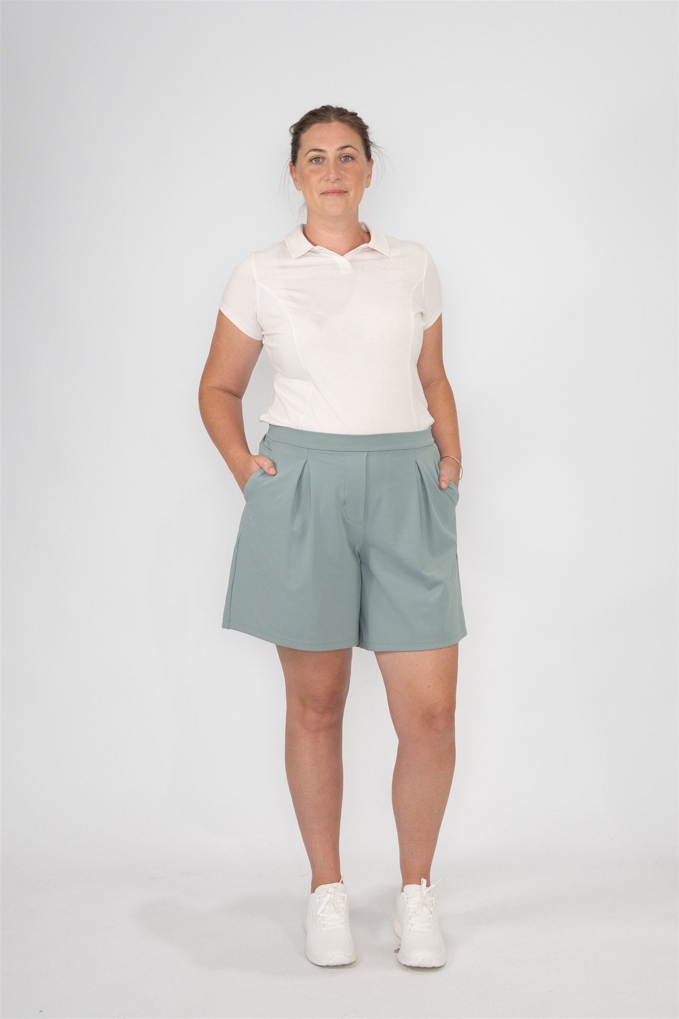 Mid-sized woman wearing Sand short sleeve polo and Fairway Green pleated golf shorts, standing front-facing with hands in pockets against a studio backdrop. Refined and versatile women’s golf apparel designed for comfort and polish.