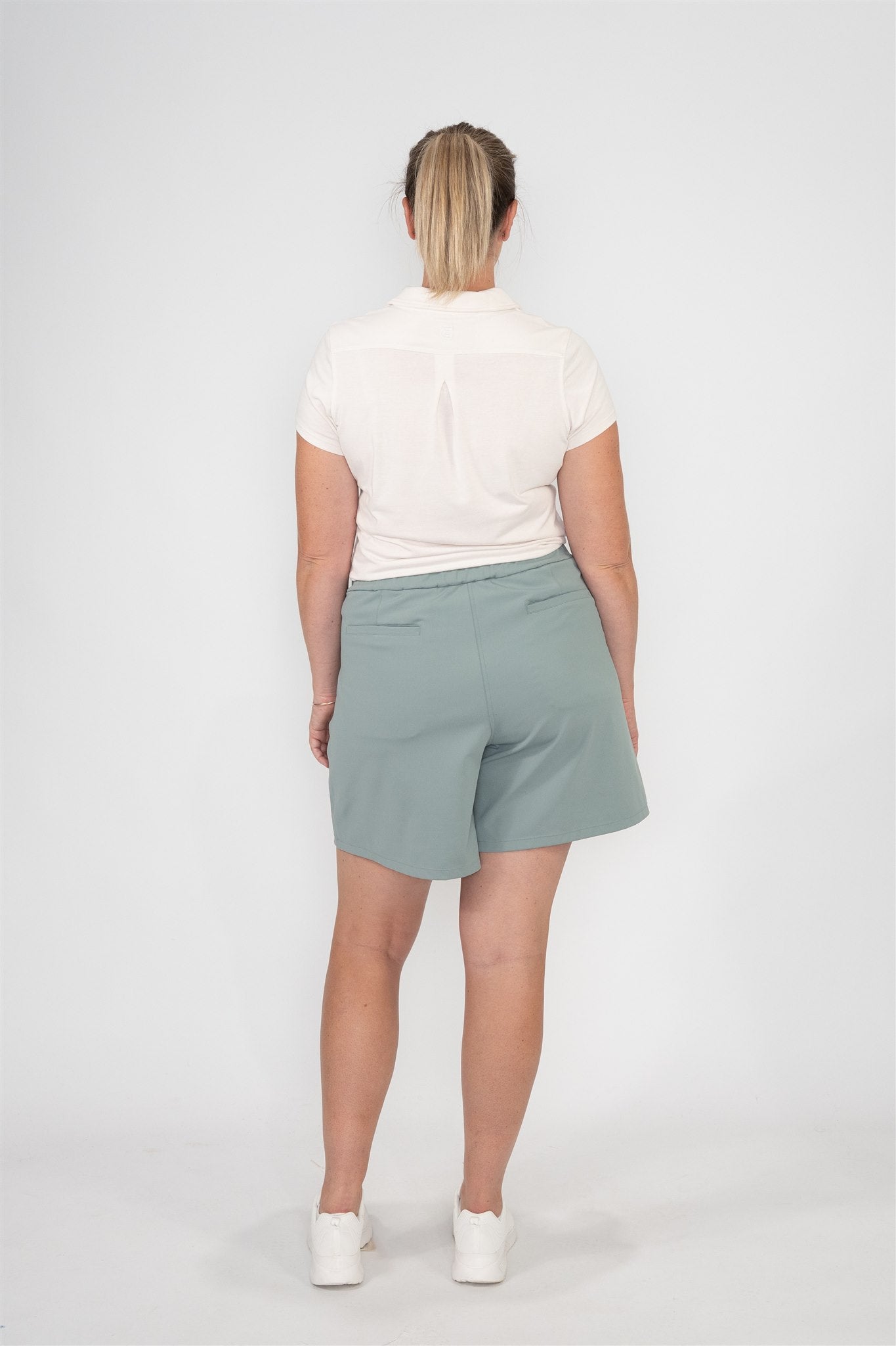 Mid-sized woman wearing Sand sleeveless polo and Fairway Green pleated golf shorts, standing in a relaxed back view pose against a white backdrop. Comfortable, tailored women’s golf apparel designed for fit, movement, and modern performance.