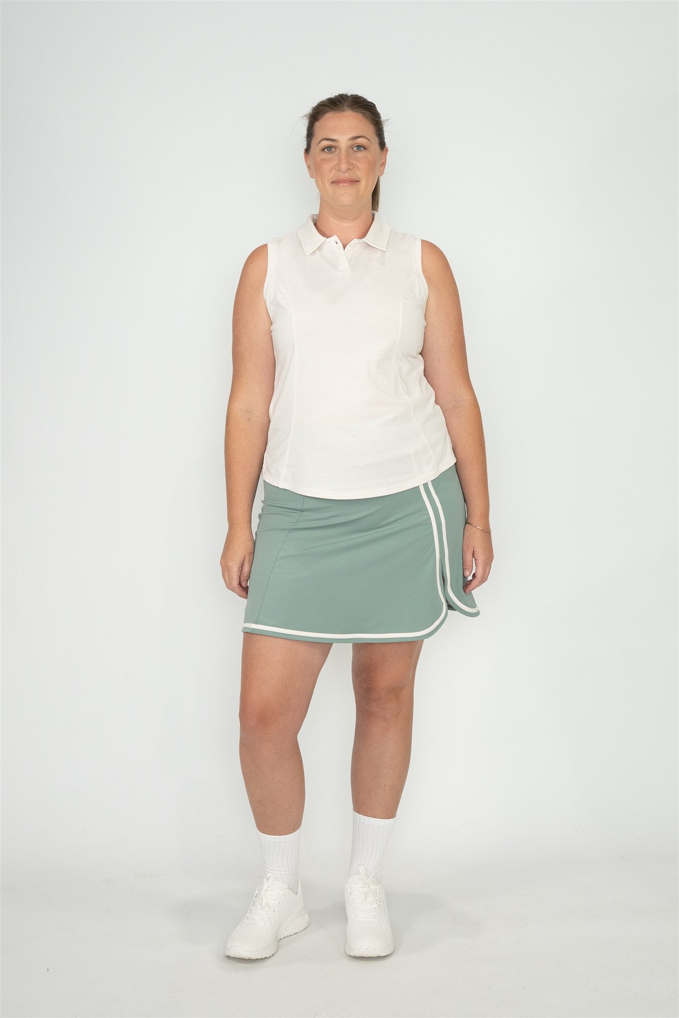 Mid size woman wearing white sleeveless polo and Fairway Green skort with white trim, standing front-facing against plain white background. Elegant and functional women’s golf apparel designed for stylish and active players of all sizes.