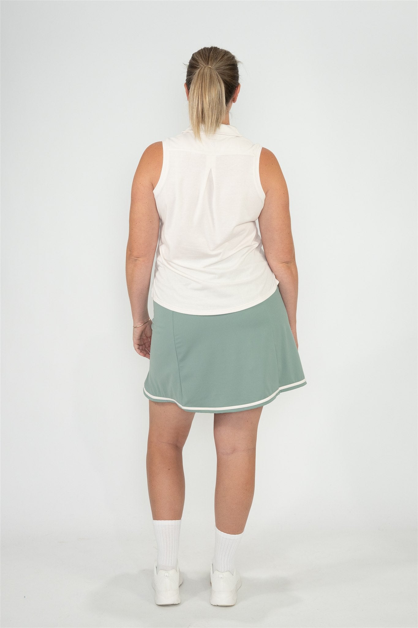 Mid-size woman wearing Sand sleeveless polo and Fairway Green wrap skort with white trim, shown from the back with arms relaxed and hair in a low ponytail. Functional and flattering women’s golf apparel, crafted for movement and all-day comfort.