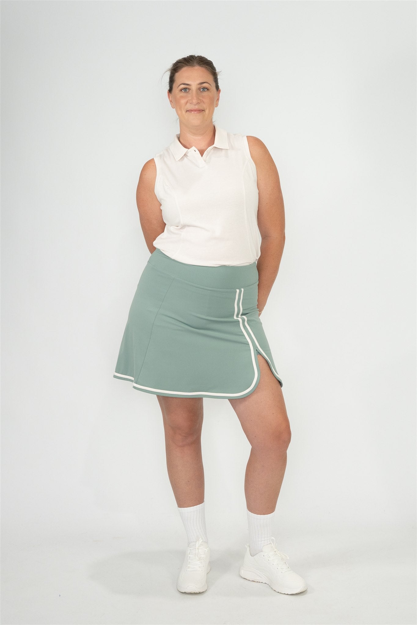 Mid-size woman wearing Sand sleeveless polo and Fairway Green wrap skort with white trim, standing front-facing with hands behind back against a clean background. Stylish and inclusive women’s golf wear designed for comfort, confidence, and performance on and off the course.
