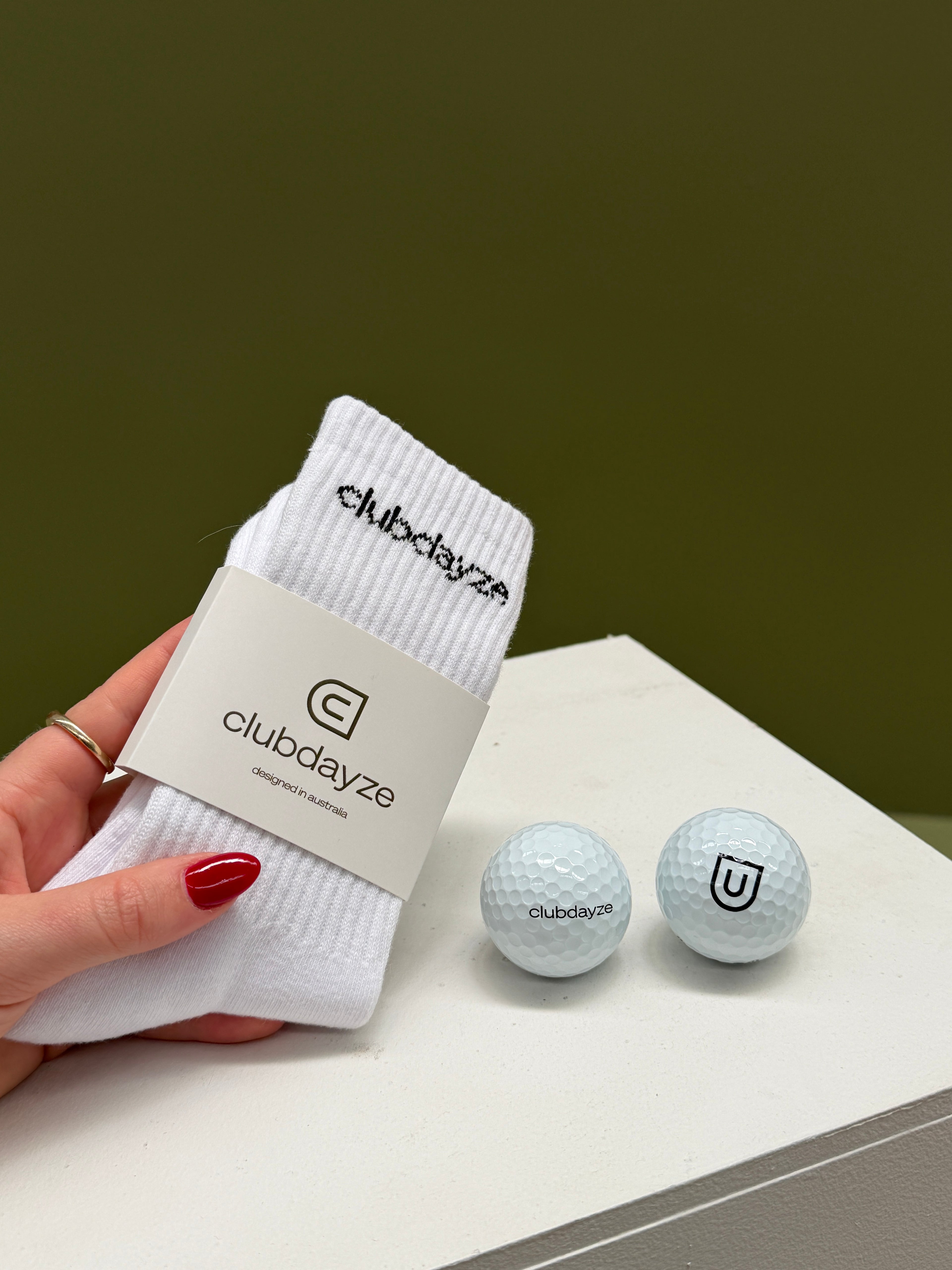 White socks with 'clubdayze' branding pack of 1 pair held by a hand, with two golf balls on a white surface.