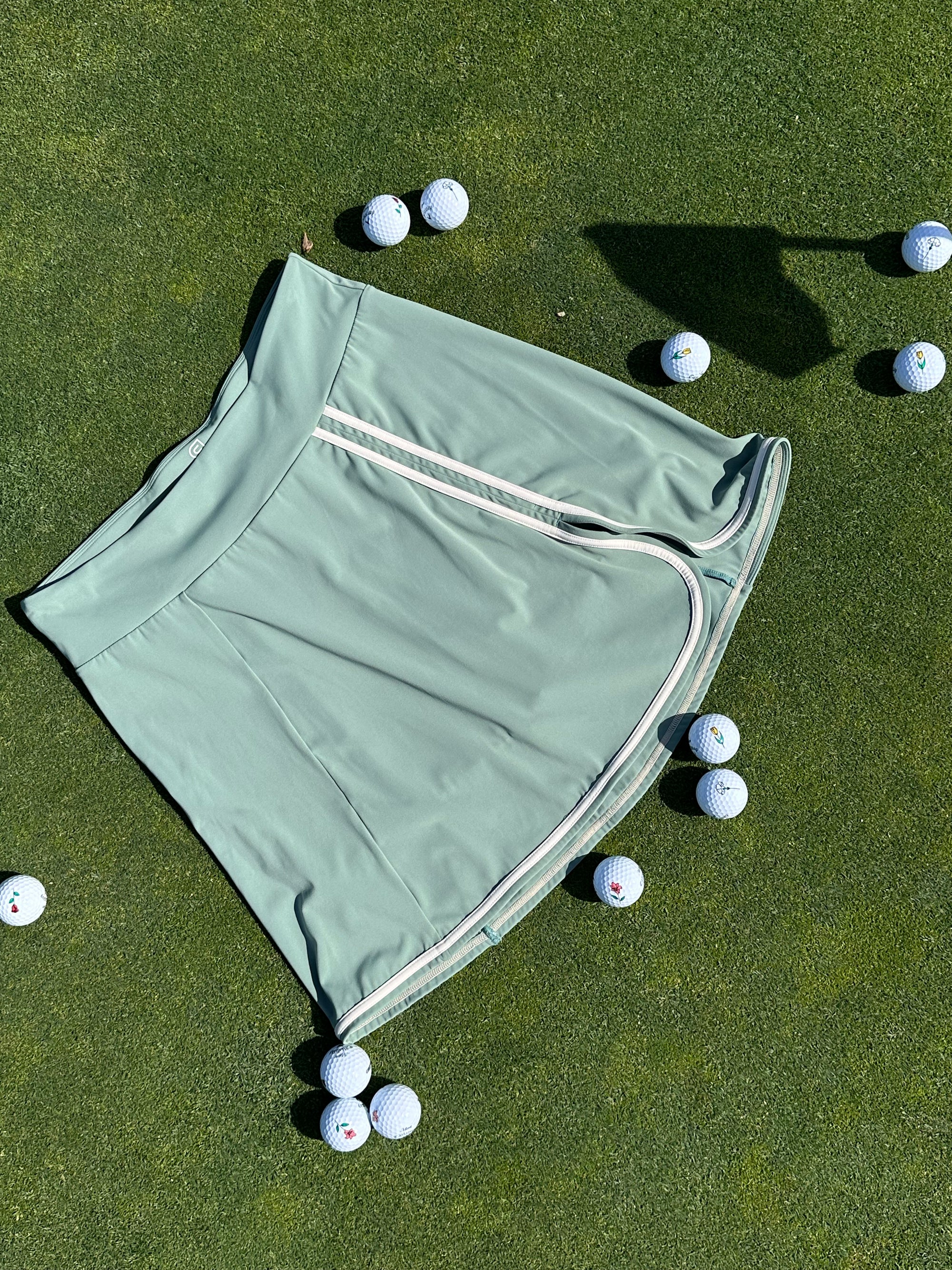 Flat lay of Fairway Green skort with Sand trim, styled on a putting green and surrounded by golf balls. Clean lines and contrast piping highlight the skort’s elegant and athletic design. Functional women’s golf wear made for movement and style.