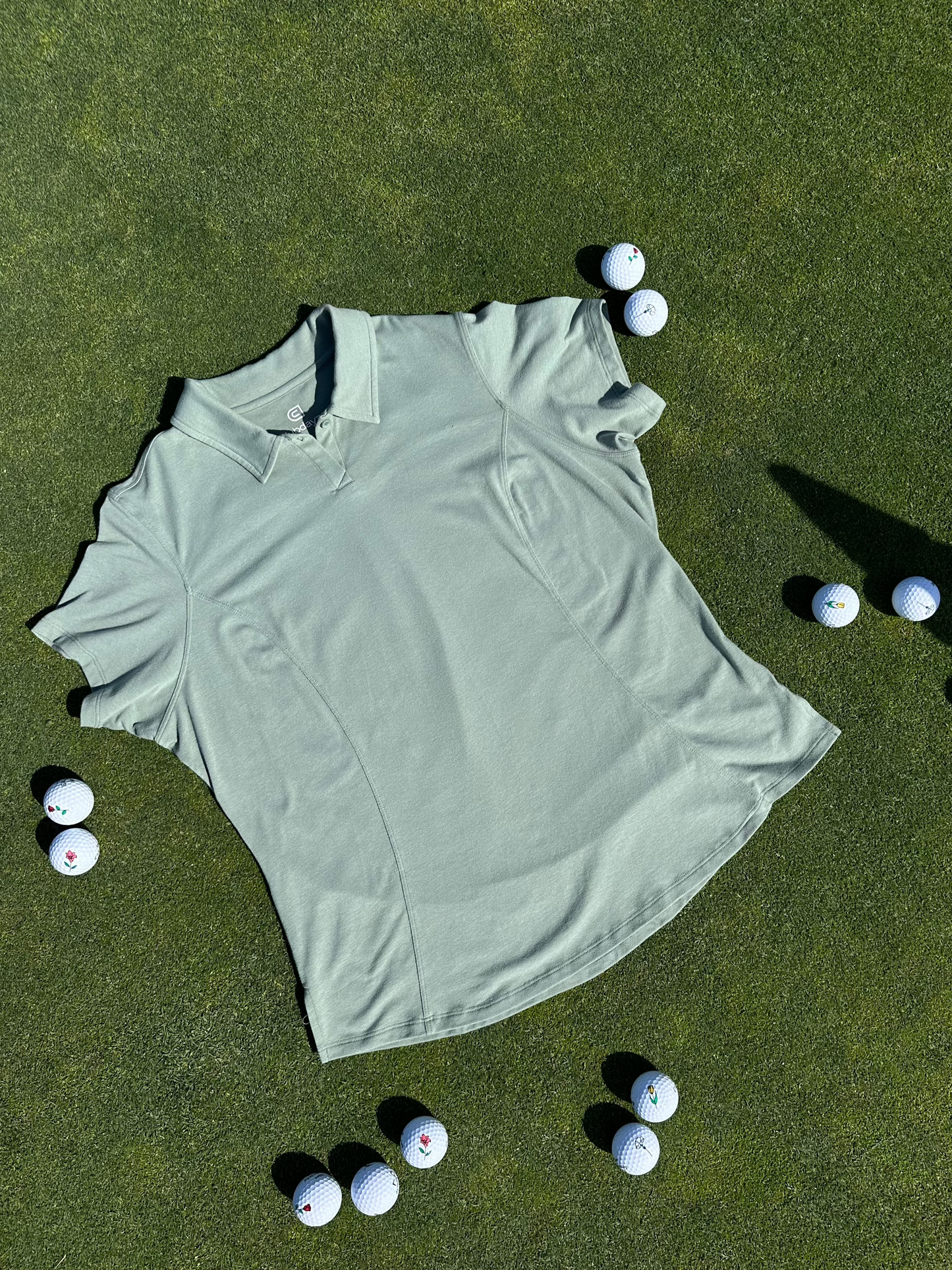 Fairway Green short sleeve women’s golf polo laid flat on grass, surrounded by golf balls. Premium, breathable fabric with elegant seam detailing designed for stylish performance on the course.