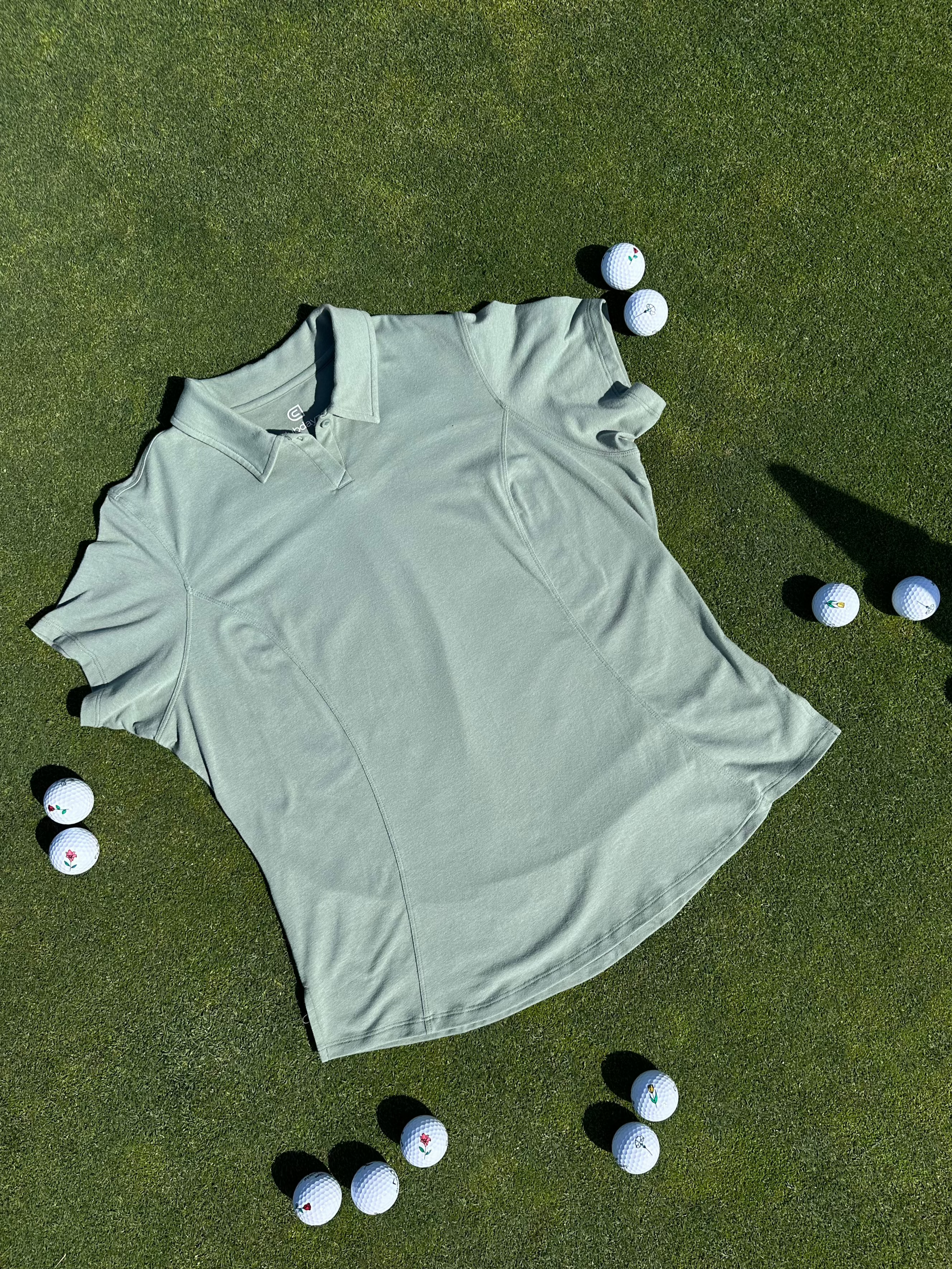 Fairway Green short sleeve women’s golf polo laid flat on grass, surrounded by golf balls. Premium, breathable fabric with elegant seam detailing designed for stylish performance on the course.