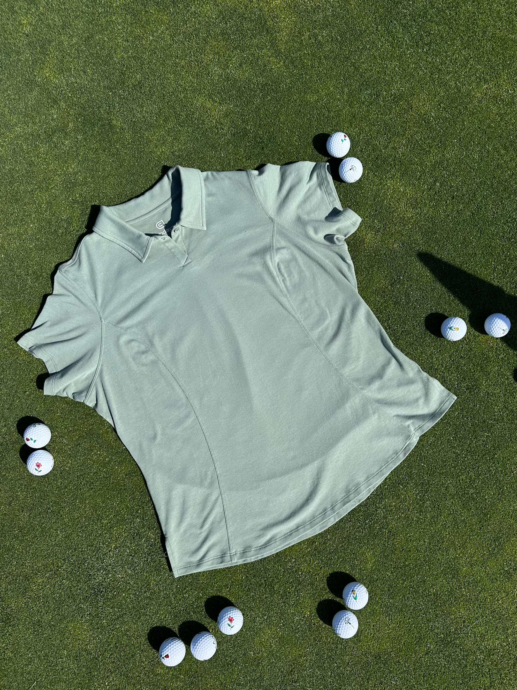 Fairway Green short sleeve women’s golf polo laid flat on grass, surrounded by golf balls. Premium, breathable fabric with elegant seam detailing designed for stylish performance on the course.