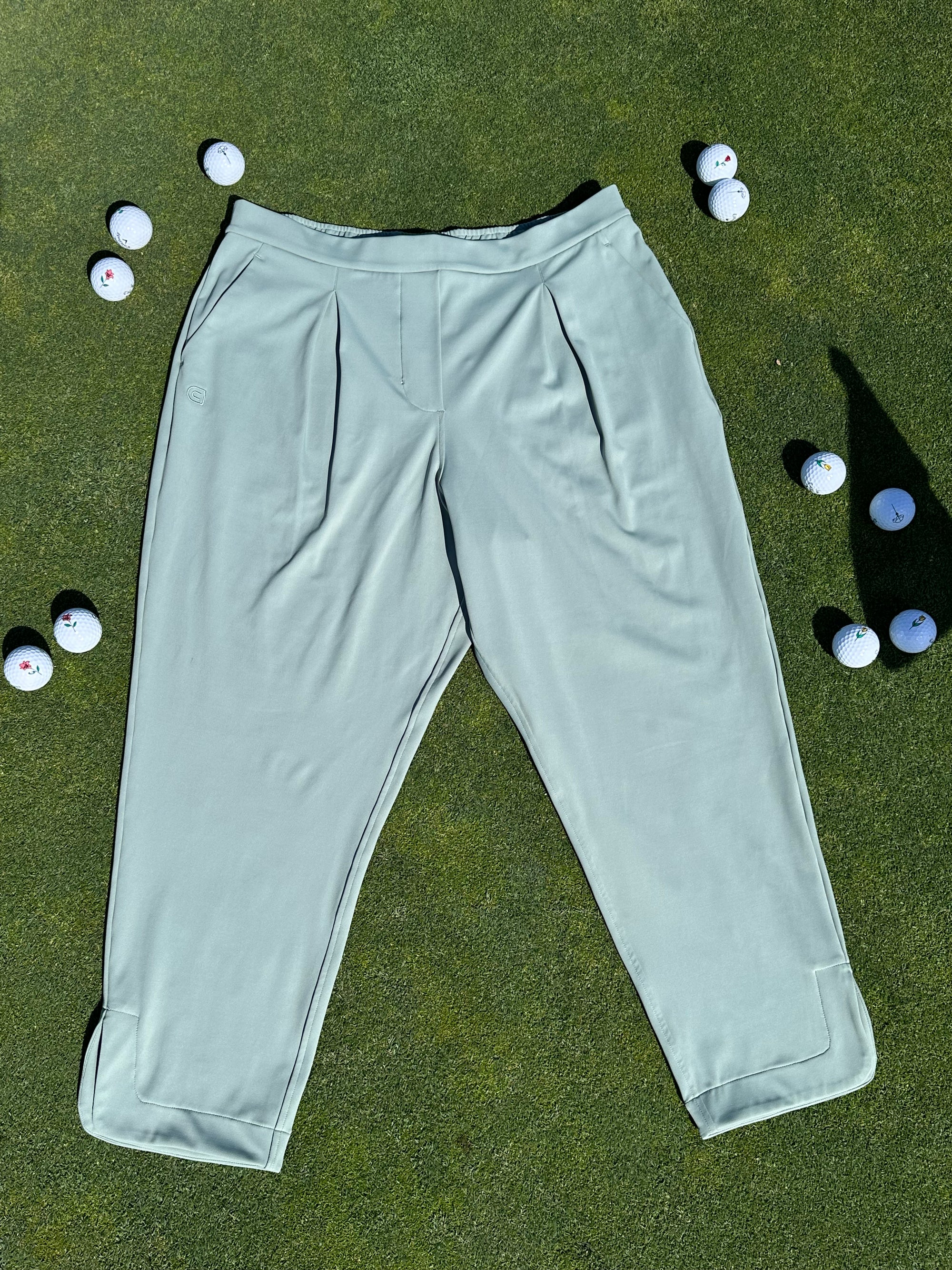 Fairway Green ankle-length golf pants laid flat on the green surrounded by scattered golf balls. Elegant and functional women’s golf apparel designed for movement and style.