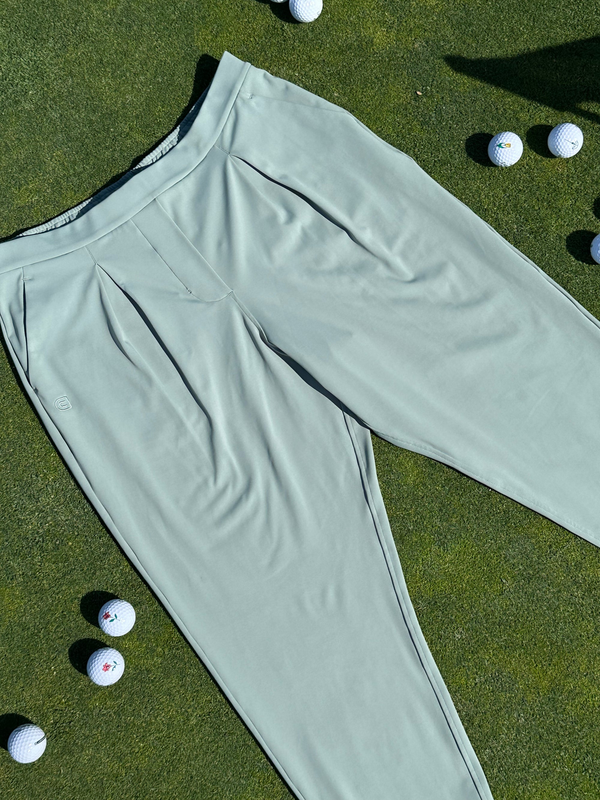 Fairway Green ankle-length golf pants with soft pleats, laid flat at an angle on a sunlit putting green, surrounded by scattered white golf balls. Sophisticated and functional women’s golf apparel designed for all-day play.