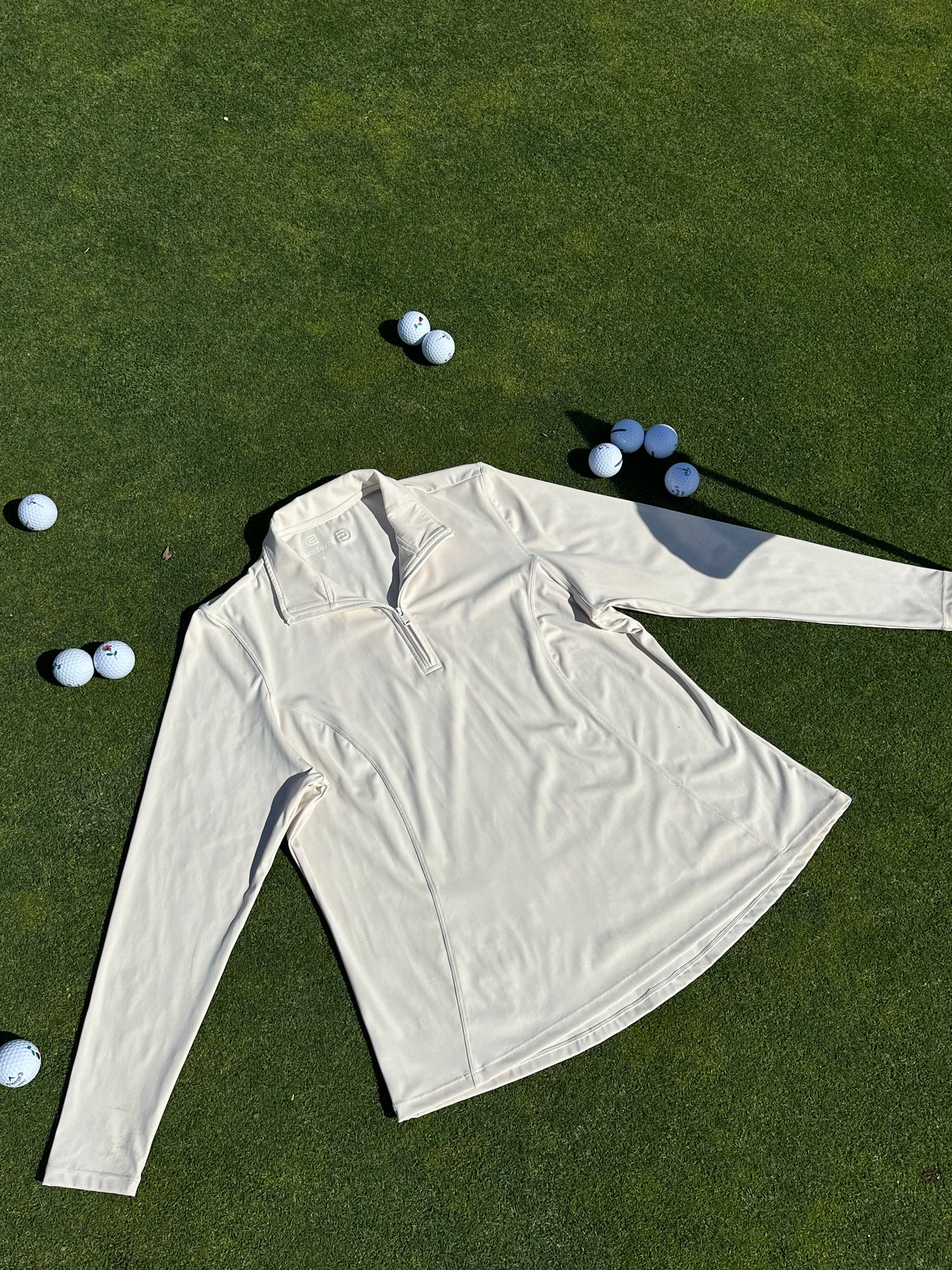 Sand quarter-zip pullover displayed flat on a sunlit green, surrounded by scattered golf balls. Lightweight and breathable women’s golf layer with sleek seam detailing and tailored silhouette, designed for performance and elegance on and off the course.