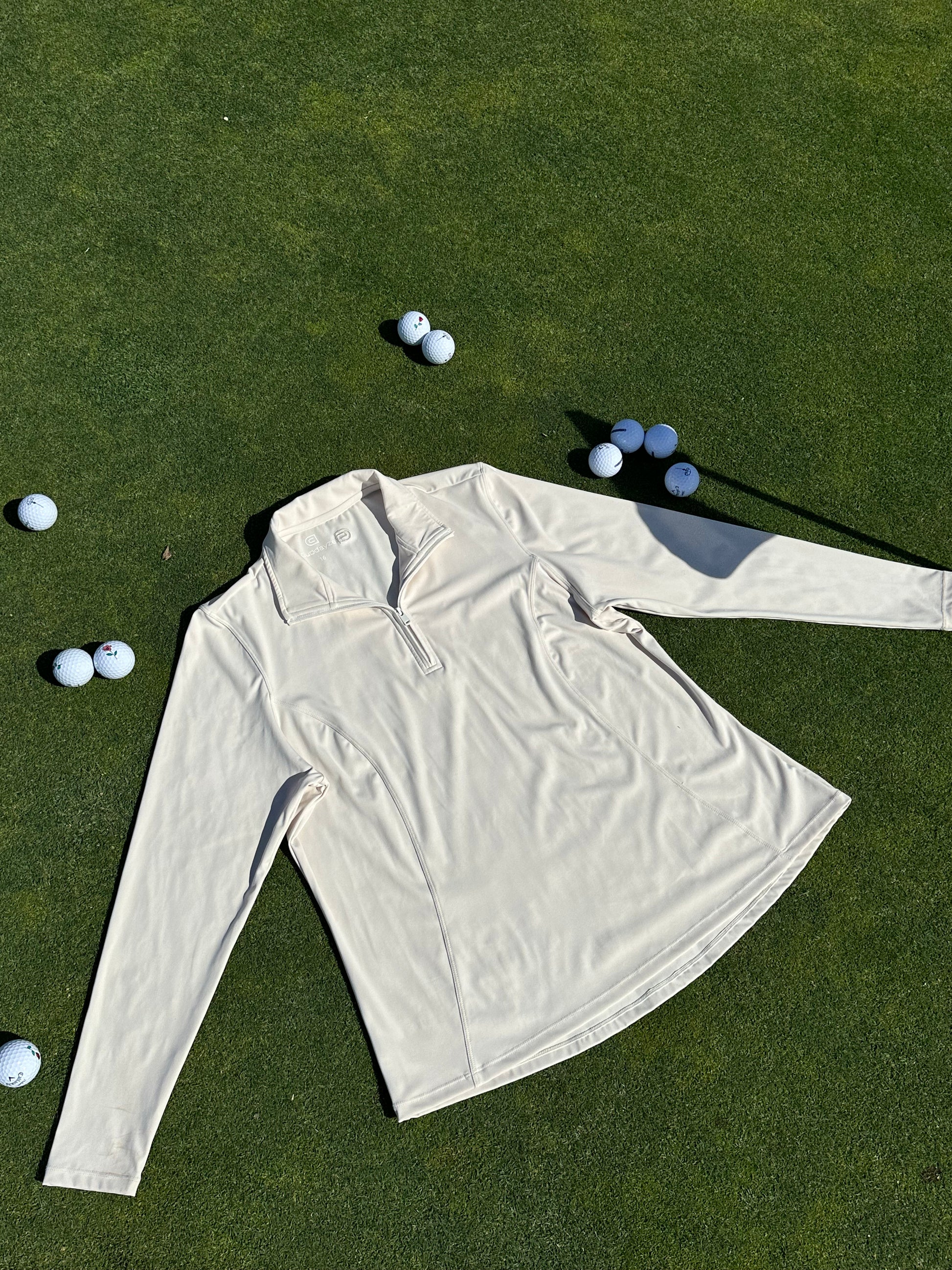 Sand quarter-zip pullover displayed flat on a sunlit green, surrounded by scattered golf balls. Lightweight and breathable women’s golf layer with sleek seam detailing and tailored silhouette, designed for performance and elegance on and off the course.