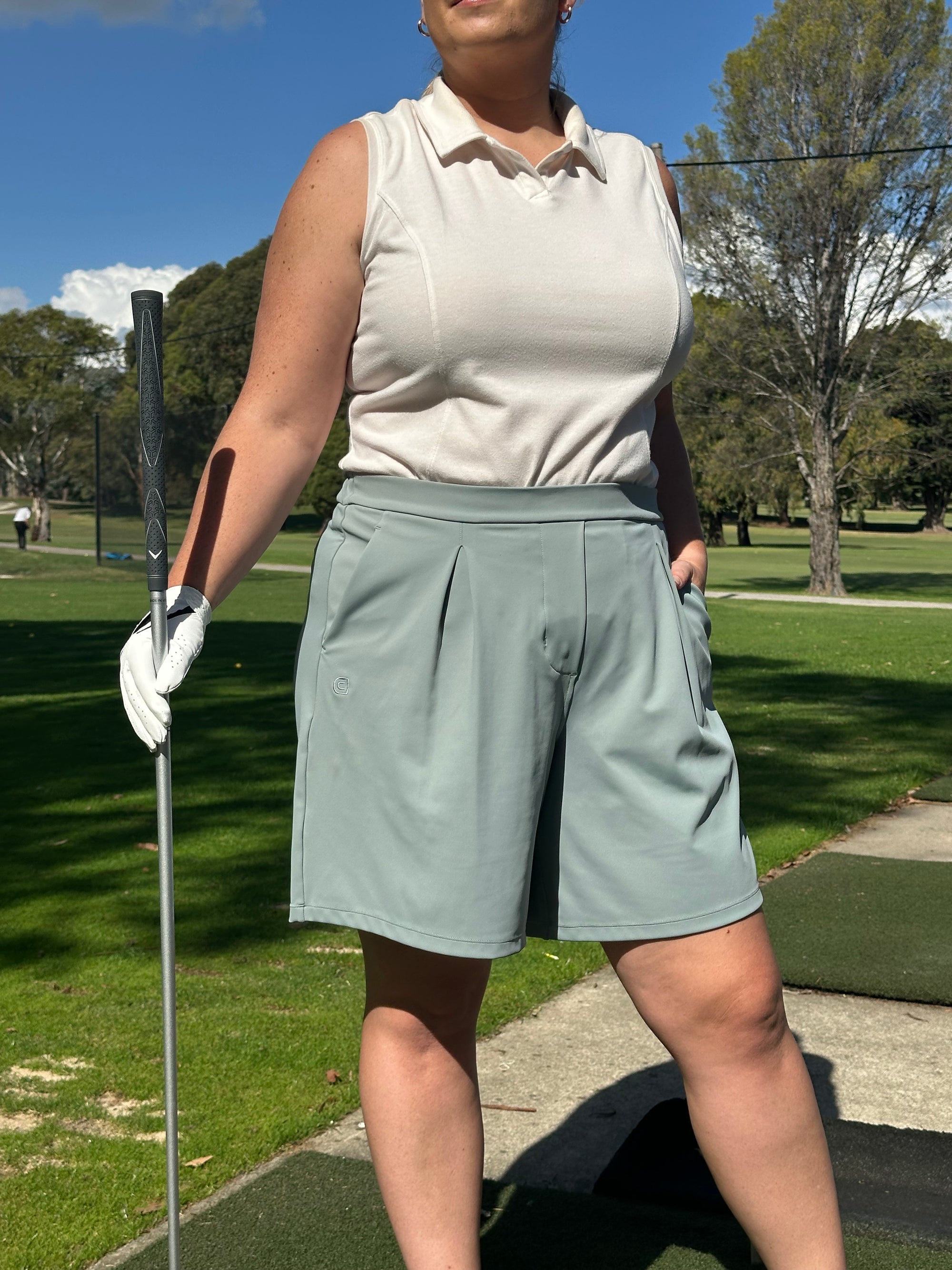 Mid-sized woman wearing Sand sleeveless polo and Fairway Green pleated golf shorts, holding a club with one hand and the other in pocket, standing confidently on a golf course. Elegant and functional women’s golf apparel for stylish and active players.