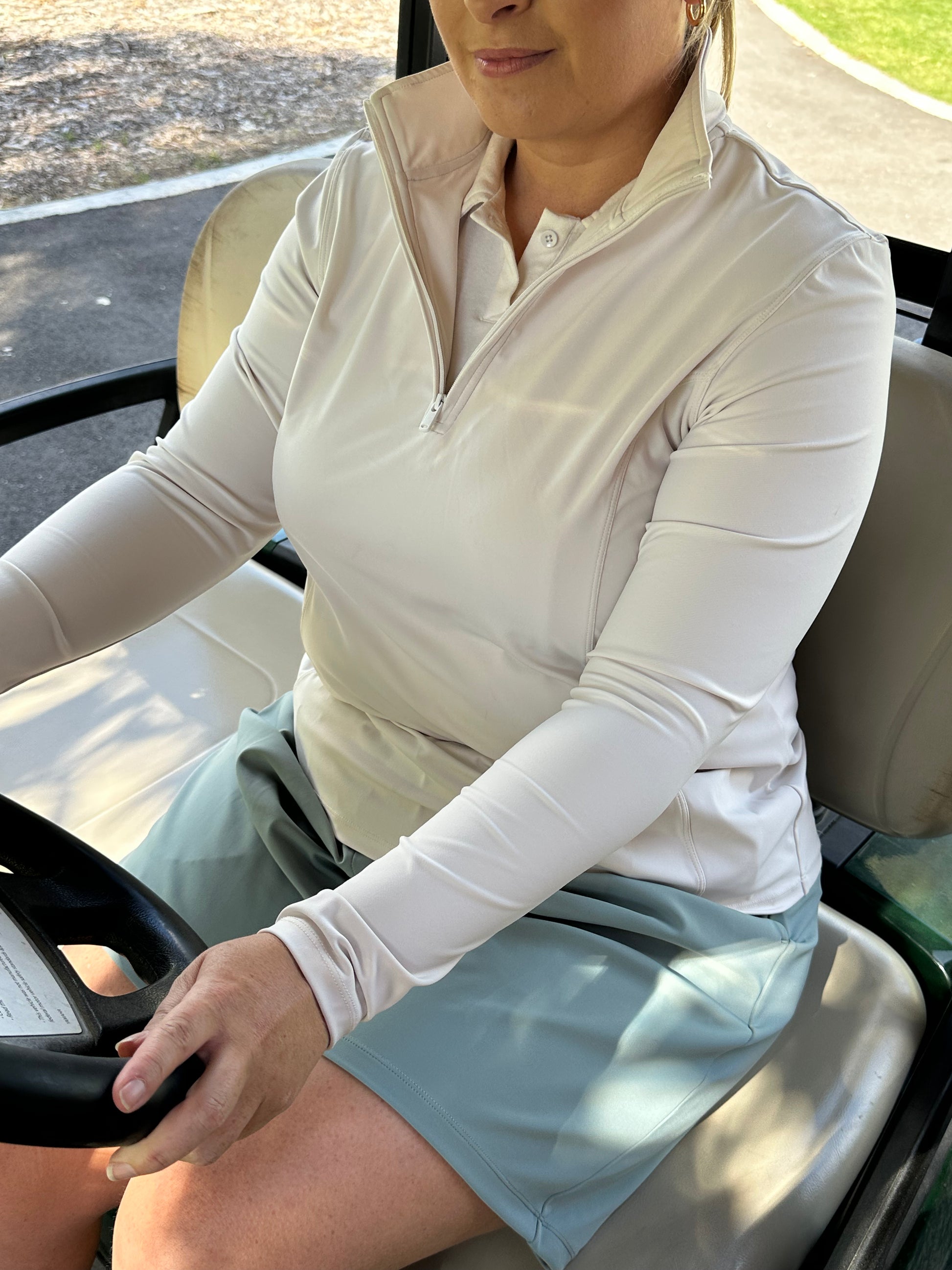 Mid-size woman seated in a golf cart wearing Sand quarter-zip pullover layered over a Sand polo and Fairway Green skort. Functional and polished women’s golf apparel designed for lightweight warmth, ease of movement, and sophisticated layering on course and in transit.
