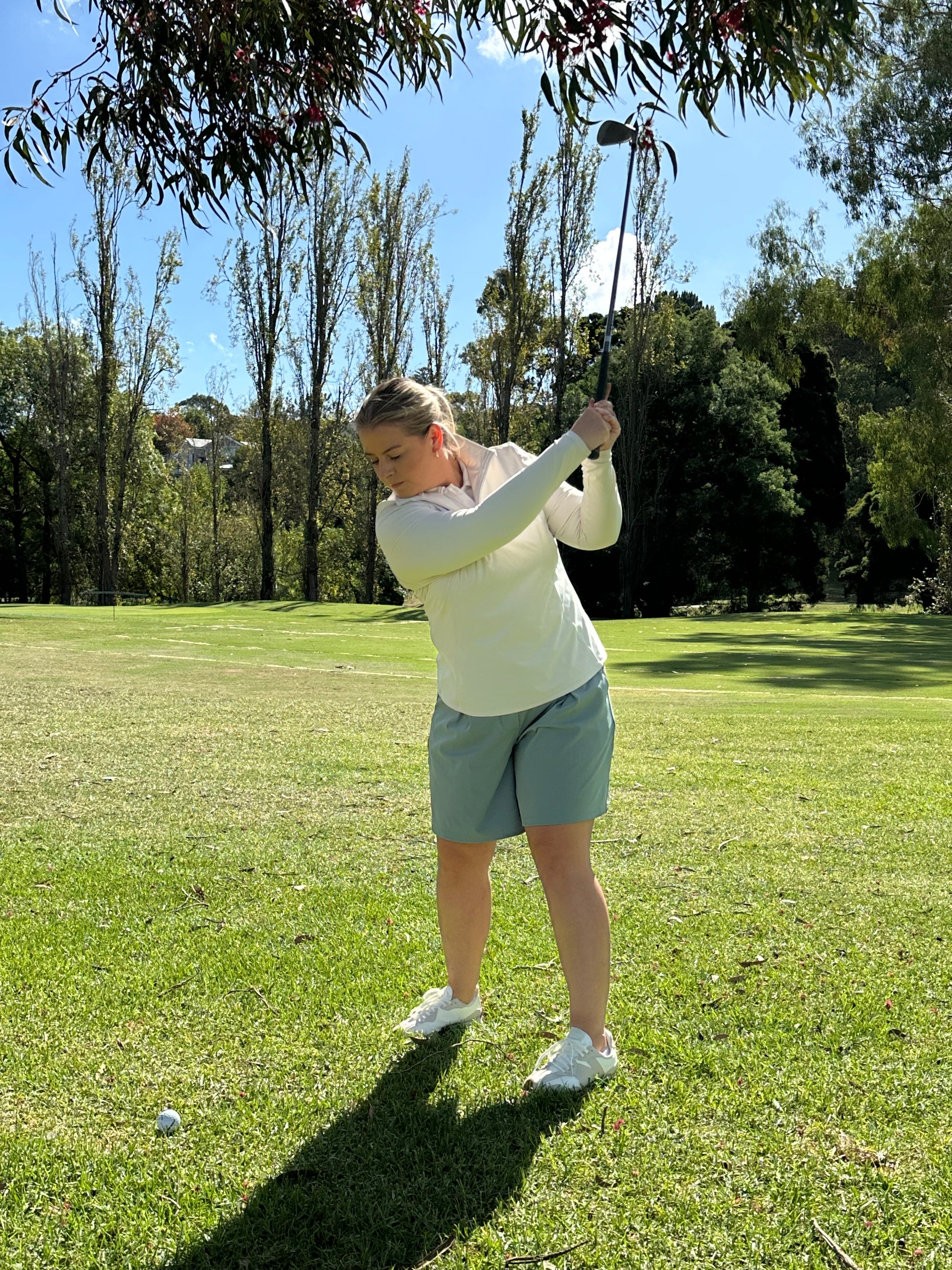 Mid-sized woman wearing Sand quarter-zip long sleeve top and Fairway Green pleated golf shorts, finishing her golf swing on a lush fairway surrounded by tall trees. Elegant and functional women’s golf wear designed for performance and sophistication.