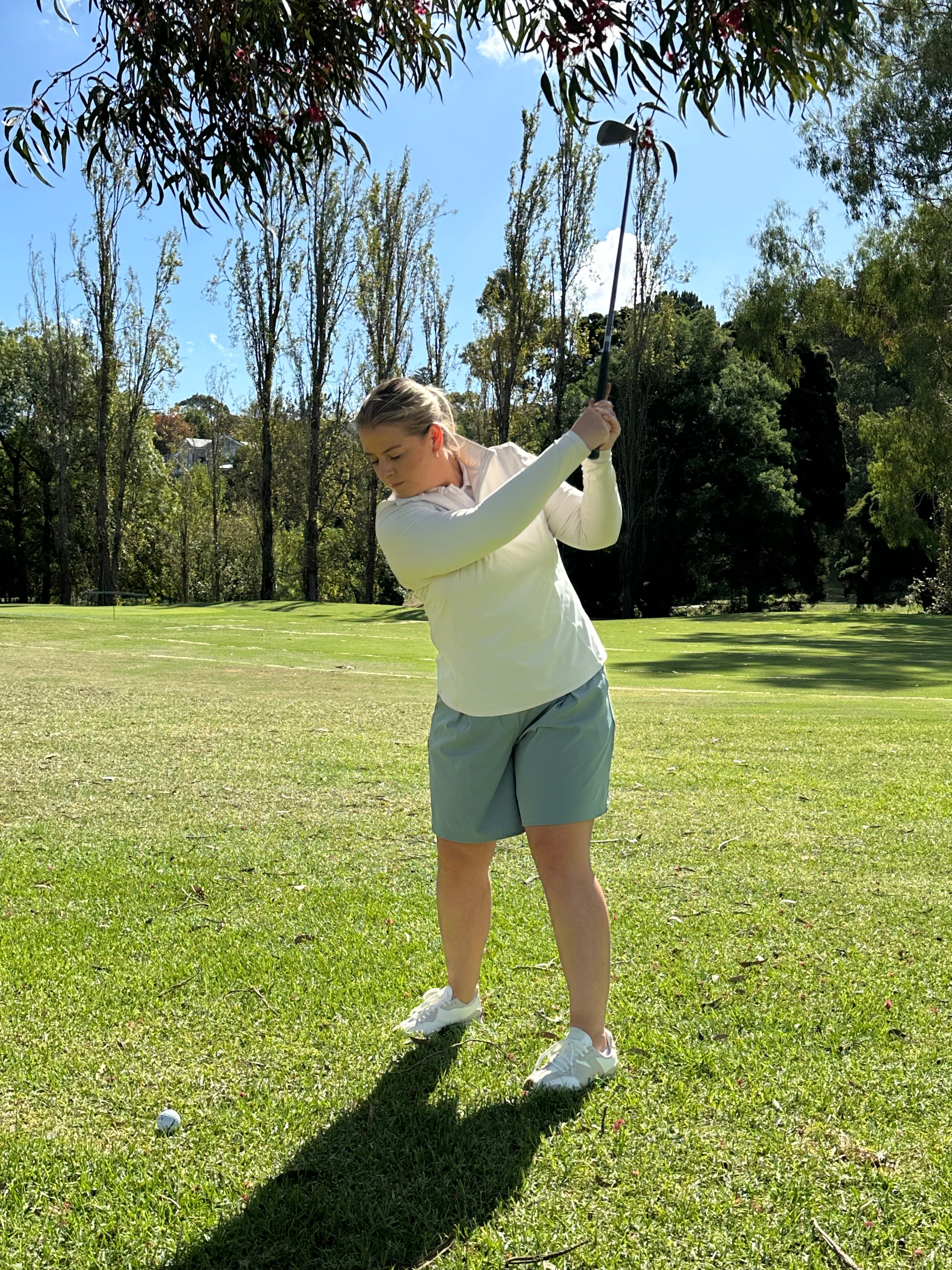 Mid-sized woman wearing Sand quarter-zip long sleeve top and Fairway Green pleated golf shorts, finishing her golf swing on a lush fairway surrounded by tall trees. Elegant and functional women’s golf wear designed for performance and sophistication.