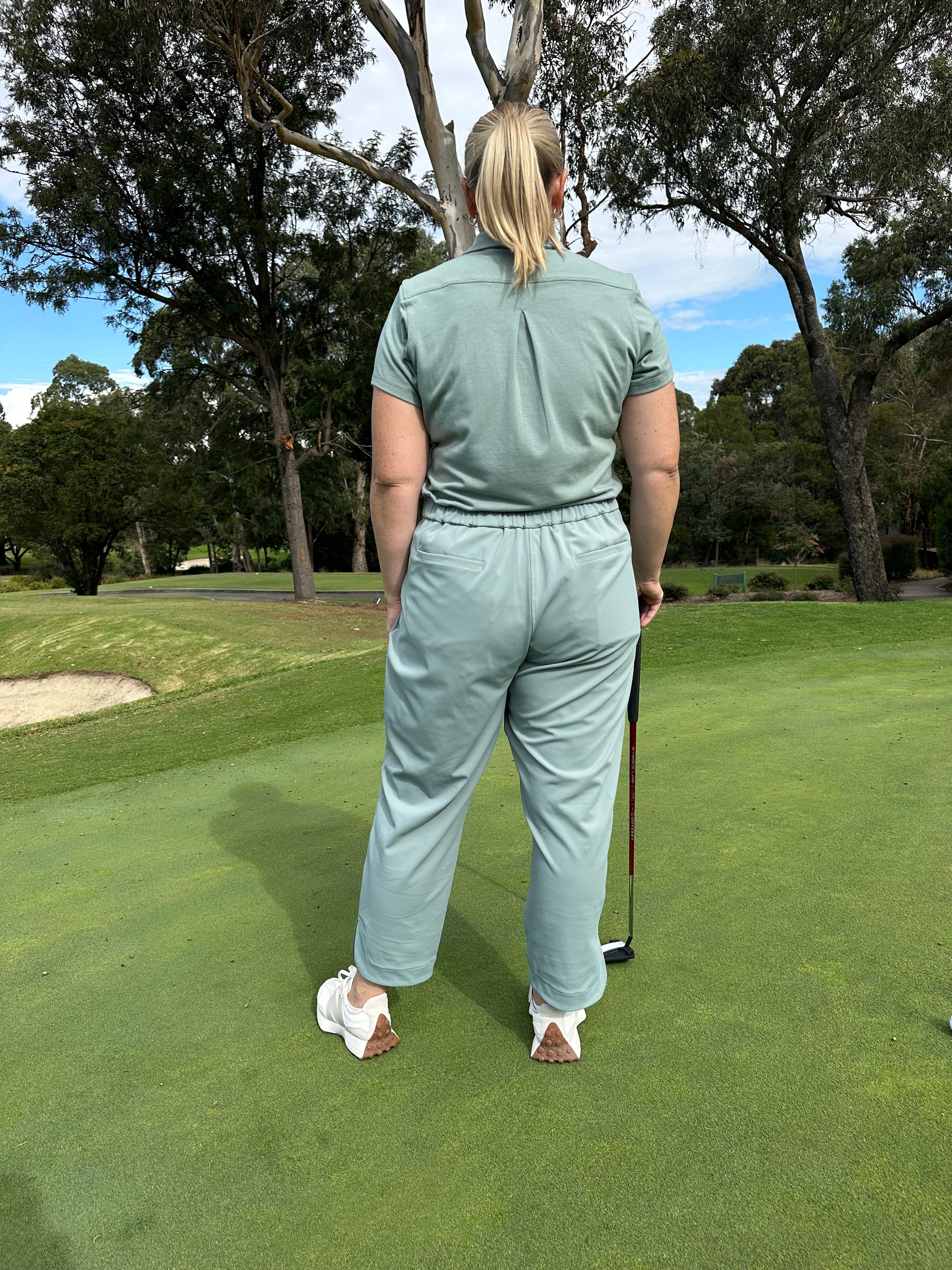 Mid-sized woman wearing Fairway Green polo and Fairway Green ankle pants, standing with back facing the camera on a putting green, holding a golf club. Premium women’s golf apparel blending style, ease, and performance.