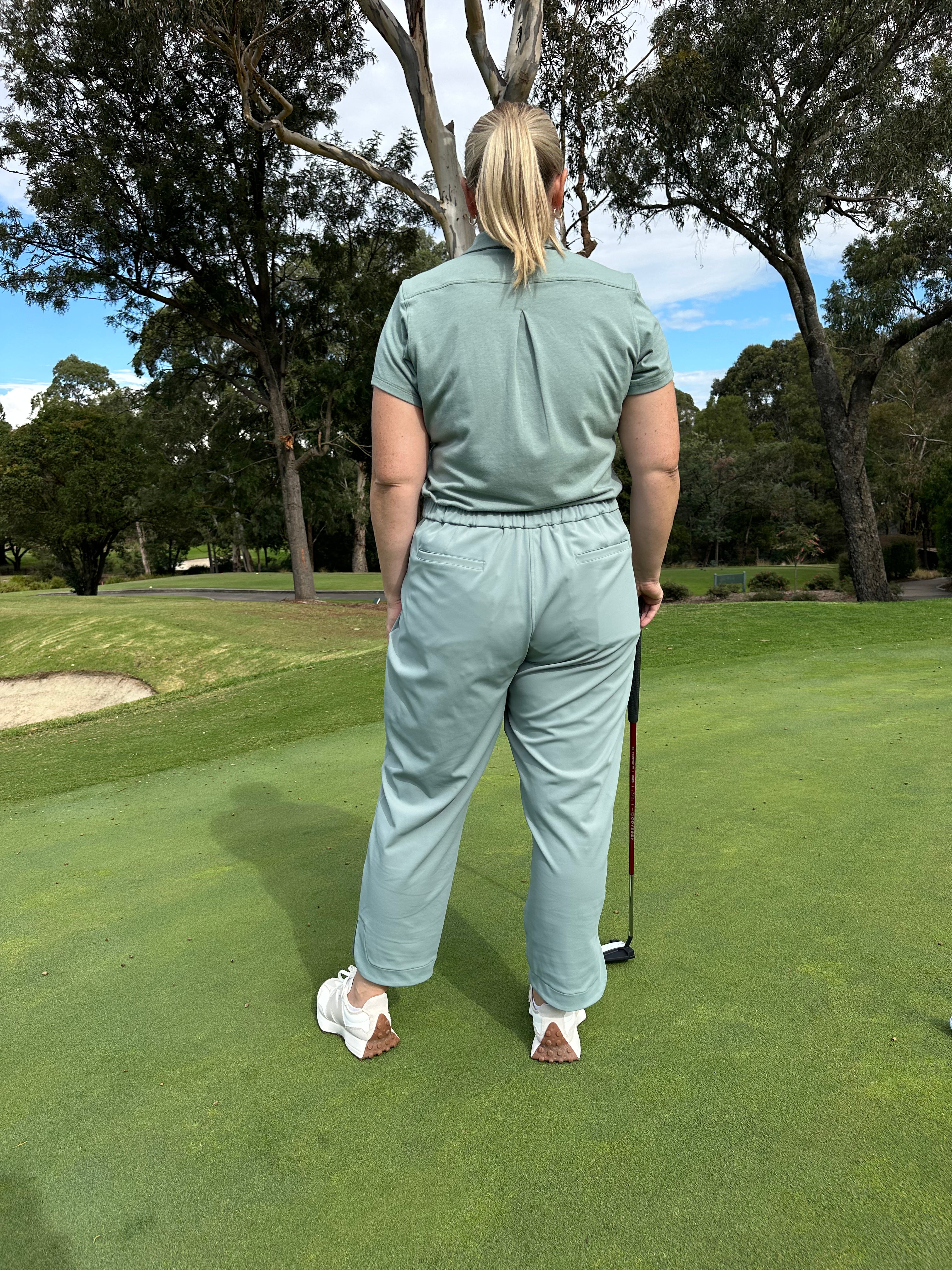 Mid-sized woman wearing Fairway Green polo and Fairway Green ankle pants, standing with back facing the camera on a putting green, holding a golf club. Premium women’s golf apparel blending style, ease, and performance.