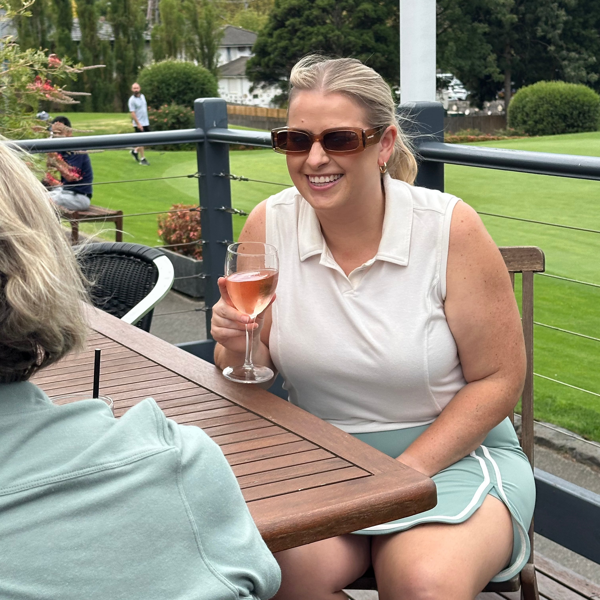 Mid-size woman wearing Sand sleeveless polo and Fairway Green wrap skort with white trim, seated outdoors with a glass of rosé, smiling in conversation. Elevated women’s golf apparel that transitions effortlessly from course to clubhouse with comfort and style.