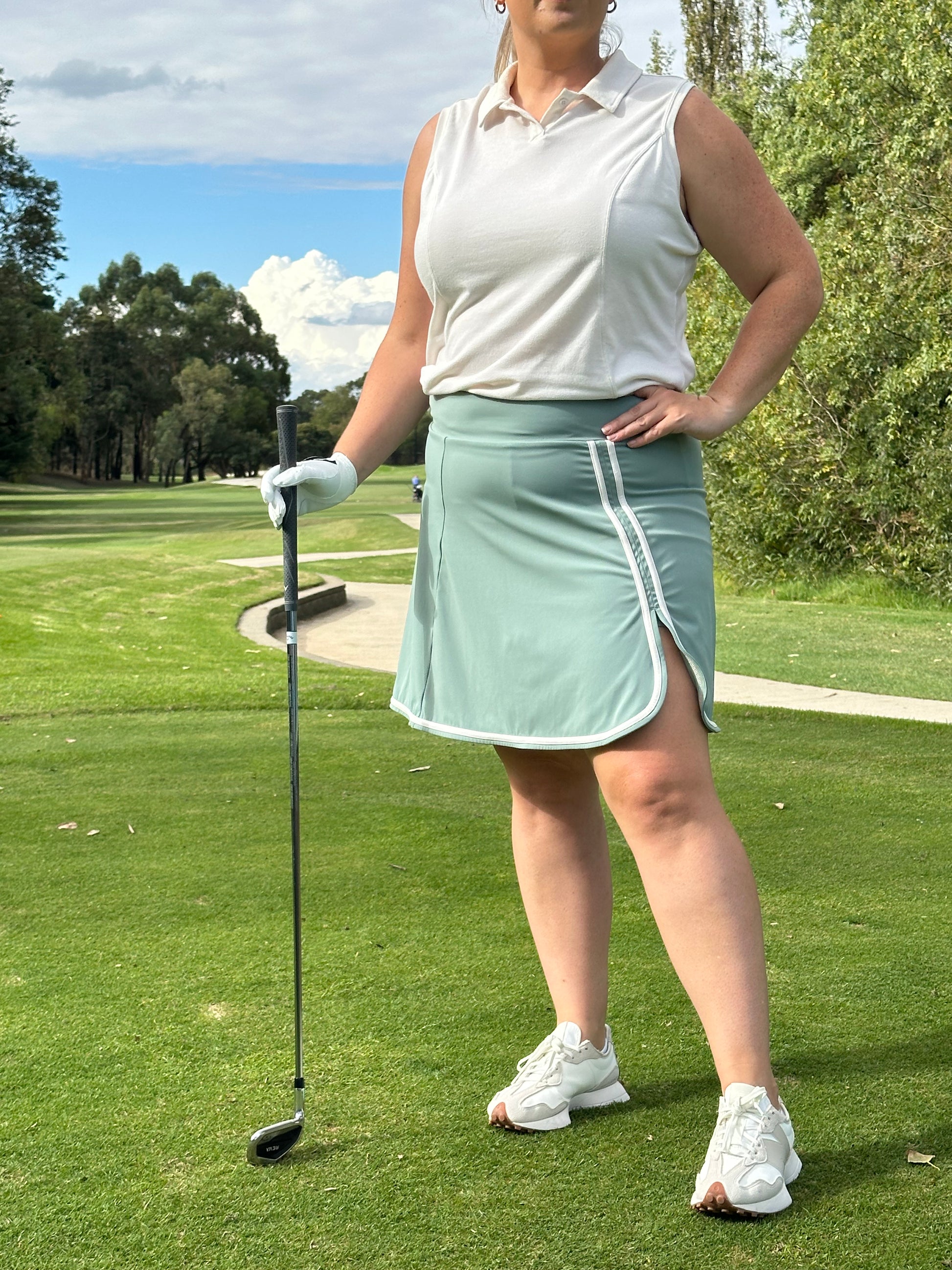 Mid-size woman wearing Sand sleeveless polo and Fairway Green wrap skort with white trim, holding a golf club and posing confidently on a sunlit course. Empowering and elegant women’s golf apparel designed for stylish performance and inclusive fit.