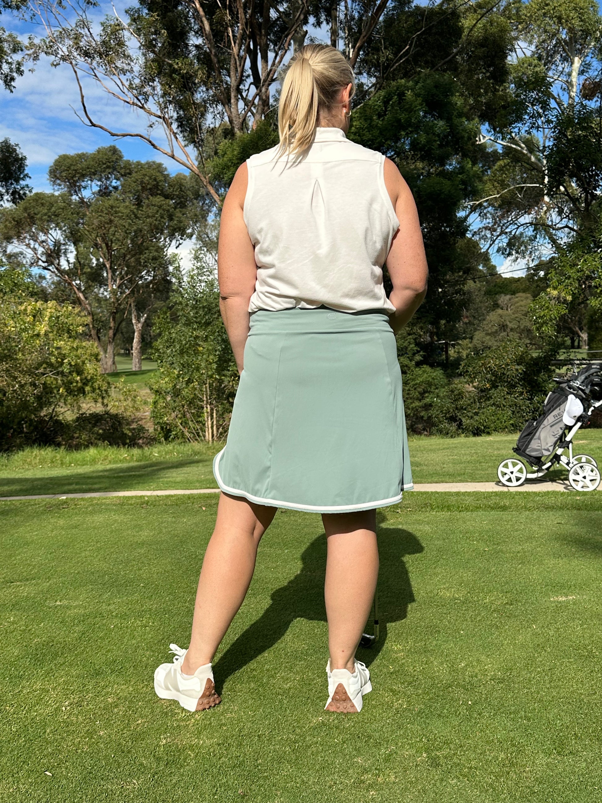 Mid-size woman wearing Sand sleeveless polo and Fairway Green wrap skort with white trim, standing with her back to the camera on a golf course. Clean, stylish women’s golf apparel crafted for movement, sun-filled days, and confident play.