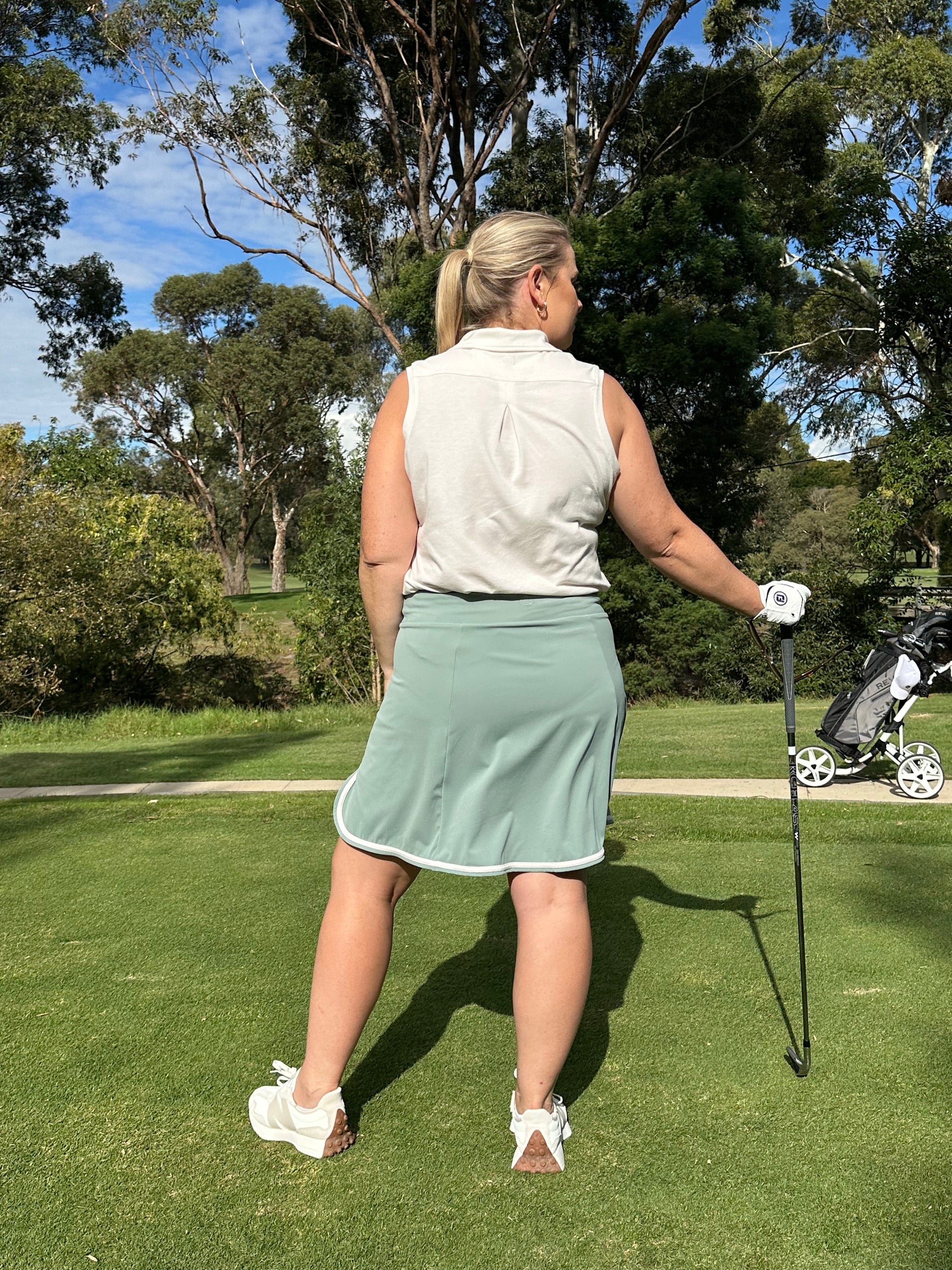 Mid-size woman wearing Sand sleeveless polo and Fairway Green wrap skort with white trim, standing on a golf course holding a club, facing away toward the fairway. Sophisticated and performance-driven women’s golf wear designed for movement, confidence, and course-ready style.