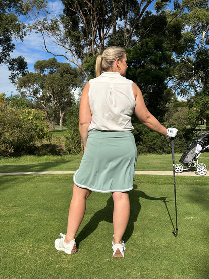 Mid-size woman wearing Sand sleeveless polo and Fairway Green wrap skort with white trim, standing on a golf course holding a club, facing away toward the fairway. Sophisticated and performance-driven women’s golf wear designed for movement, confidence, and course-ready style.