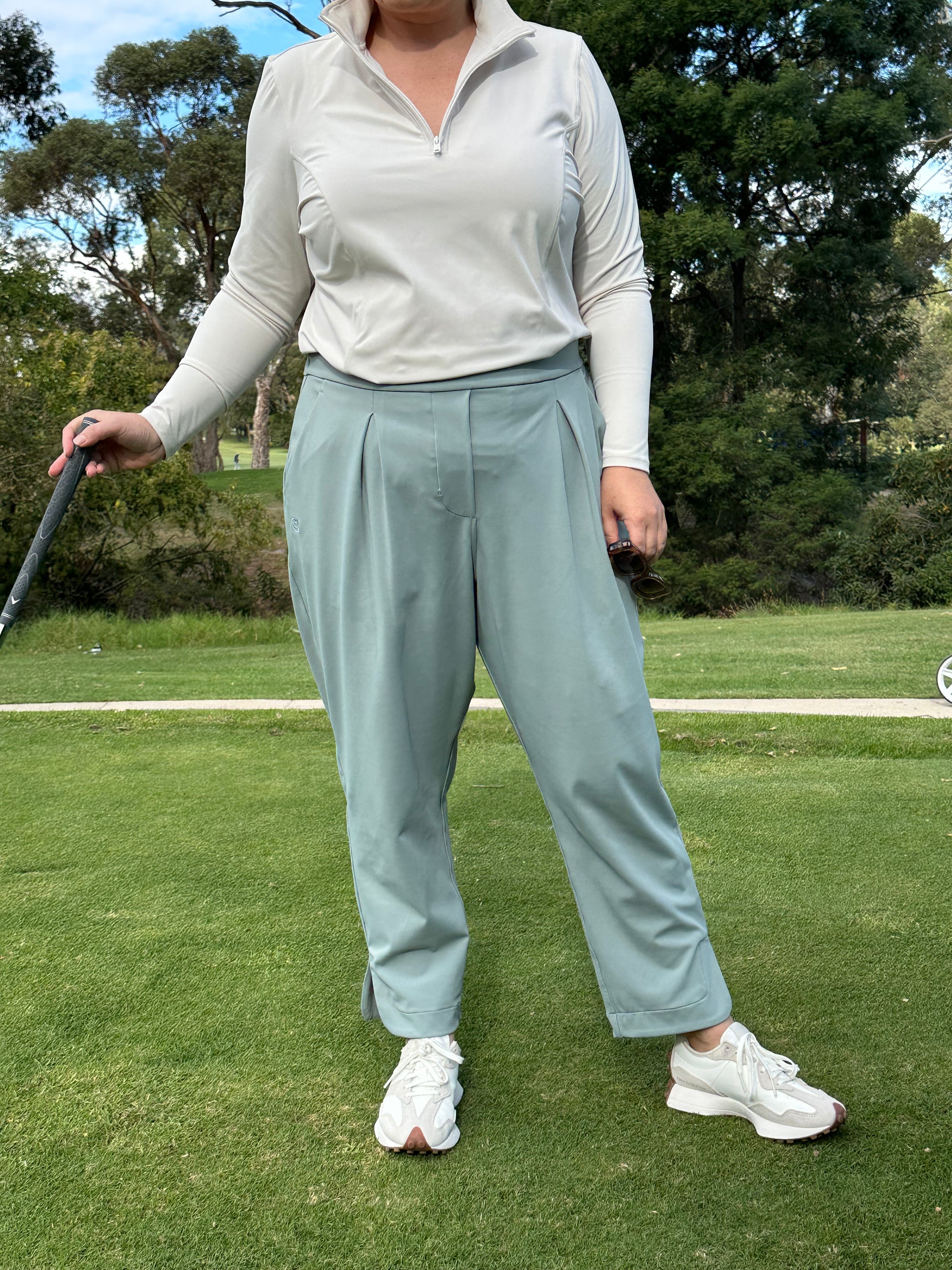 Mid-sized woman wearing Sand quarter zip and Fairway Green ankle pants, standing in address position on the golf course. Elegant and functional women’s golf wear designed for confident, active players.