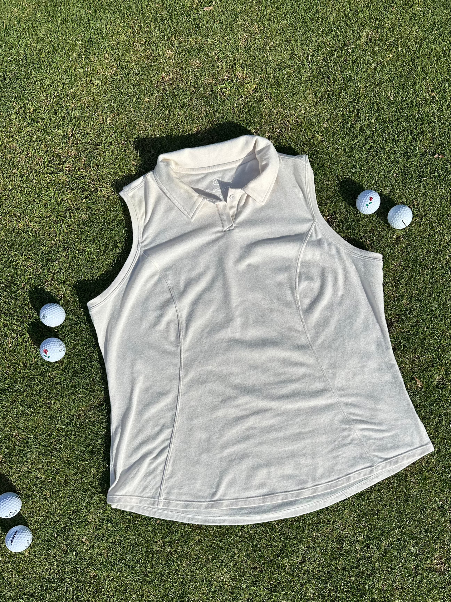 Sand sleeveless polo laid flat on vibrant green grass surrounded by scattered golf balls. Elegant and functional women’s golf apparel, perfect for stylish and active players.