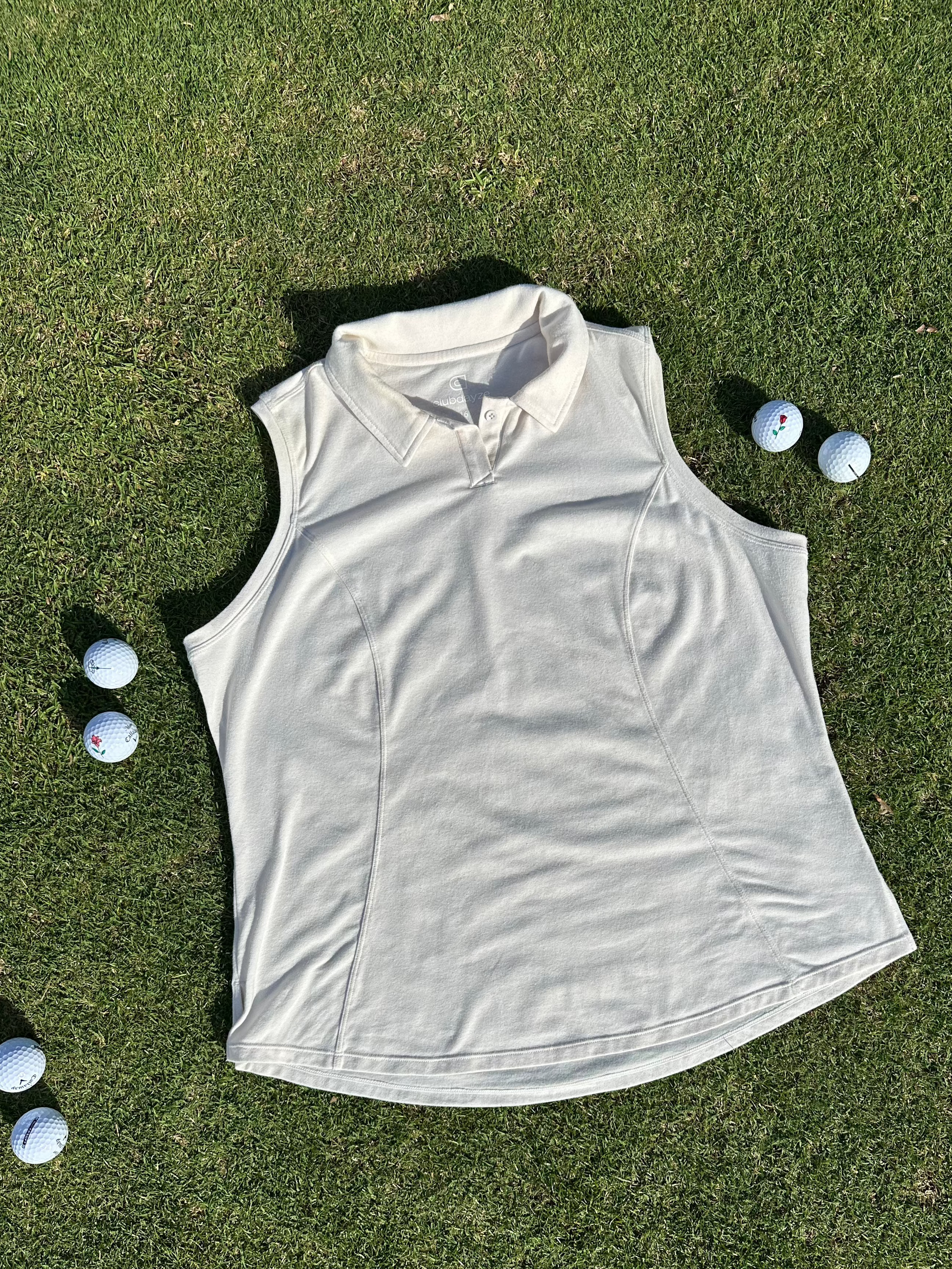 Sand sleeveless polo laid flat on vibrant green grass surrounded by scattered golf balls. Elegant and functional women’s golf apparel, perfect for stylish and active players.
