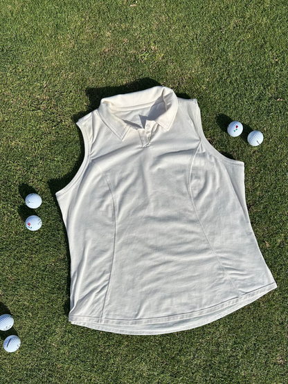 Sand sleeveless polo laid flat on vibrant green grass surrounded by scattered golf balls. Elegant and functional women’s golf apparel, perfect for stylish and active players.