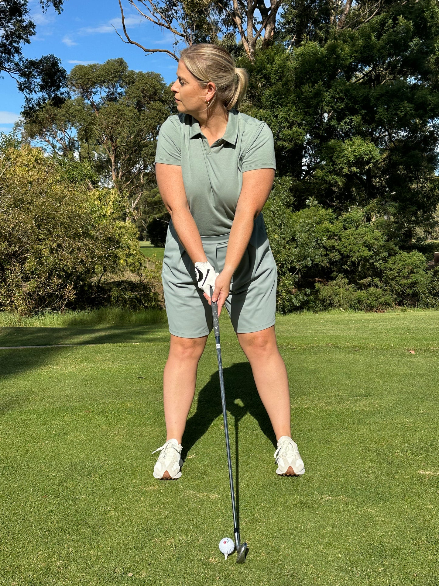 Mid-size woman wearing Fairway Green short sleeve polo and matching pleated golf shorts, preparing to swing on a sunlit course. Inclusive and performance-driven women’s golf apparel designed for focus, flexibility, and confident play.
